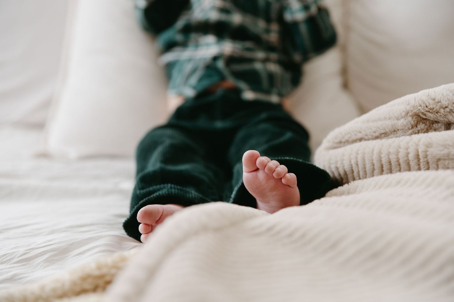 Close-up of a baby's tiny feet in dark green corduroy pants on a white bed.