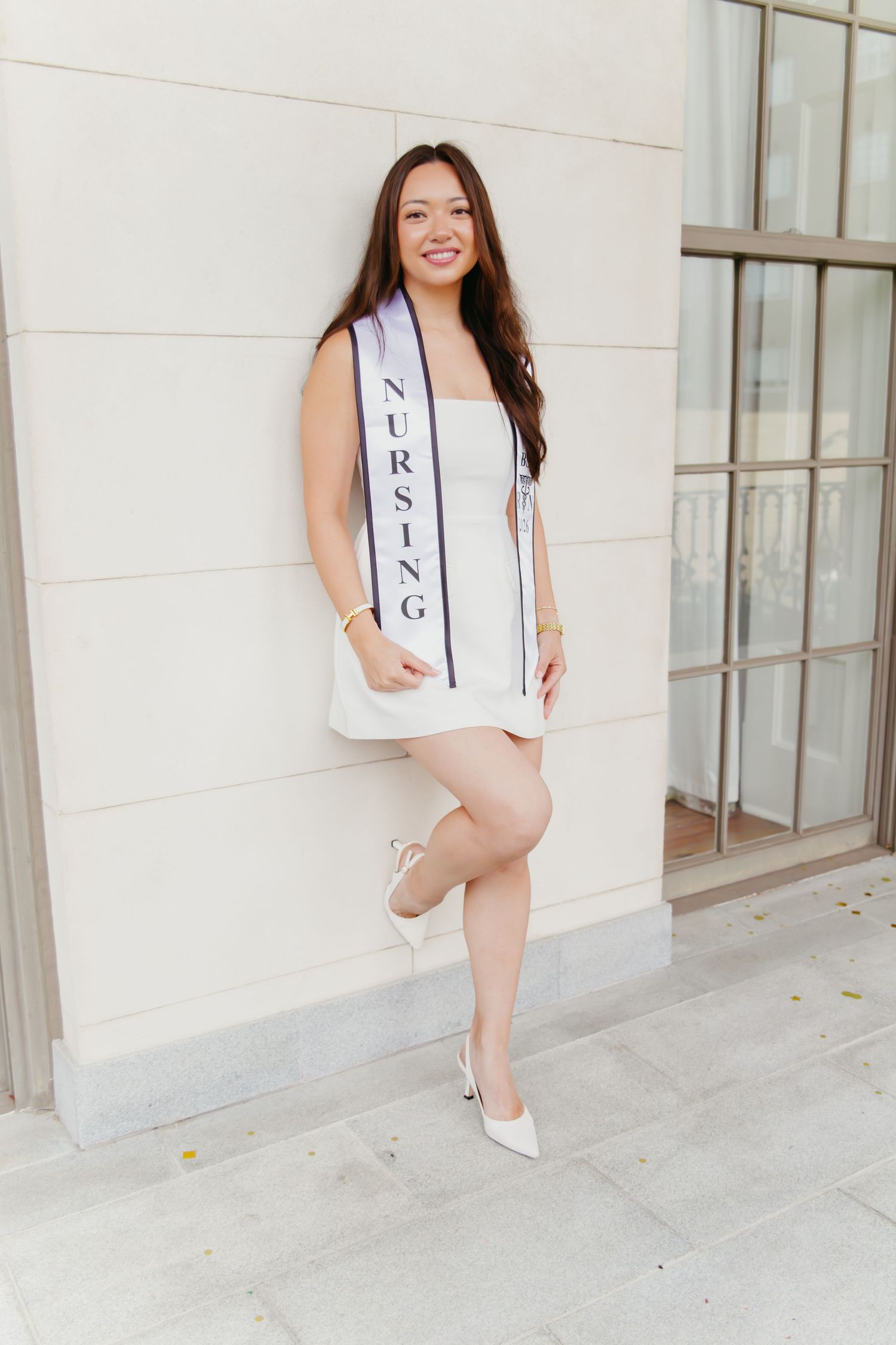 Nursing graduate in white dress and heels leaning against building wall outdoors.