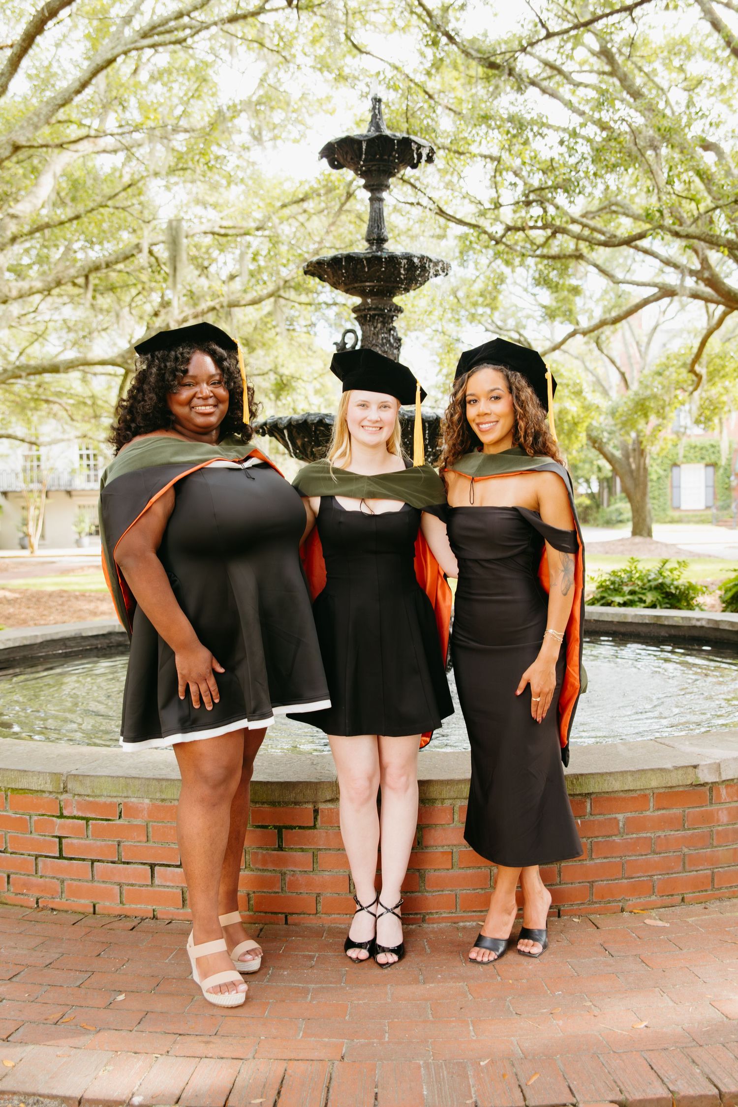 Three female graduates in black dresses and academic regalia pose by a fountain.