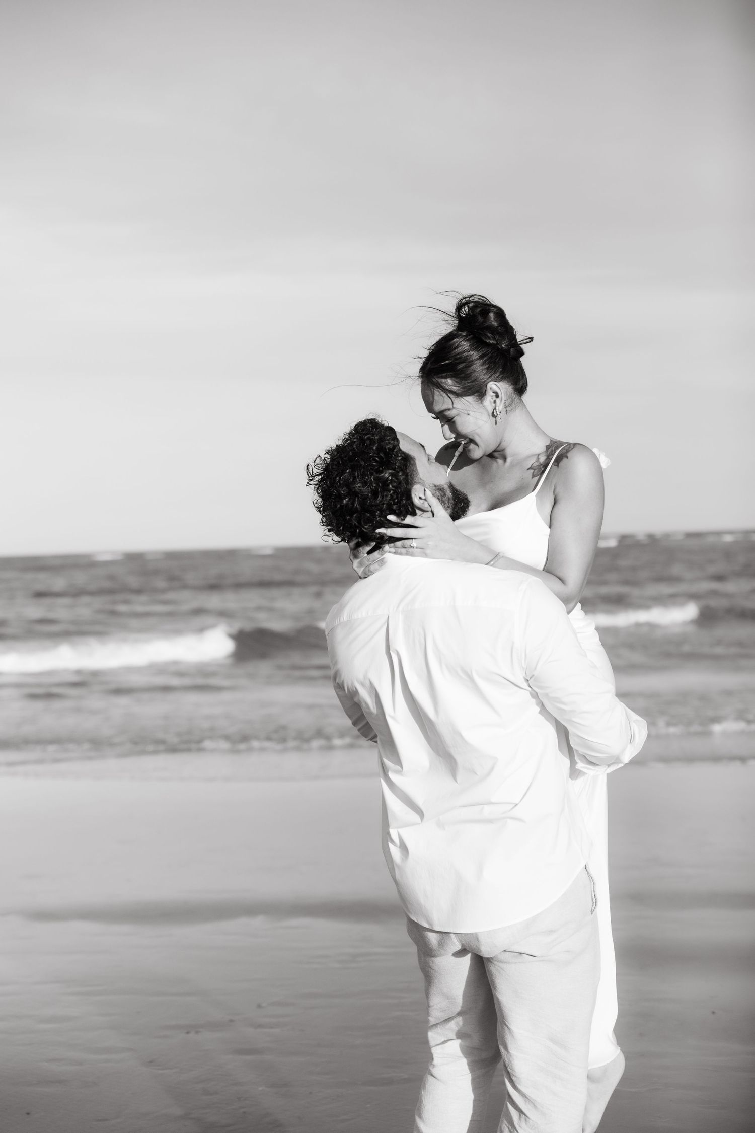 Black and white romantic photo of a couple embracing and laughing together on a beach by the ocean.