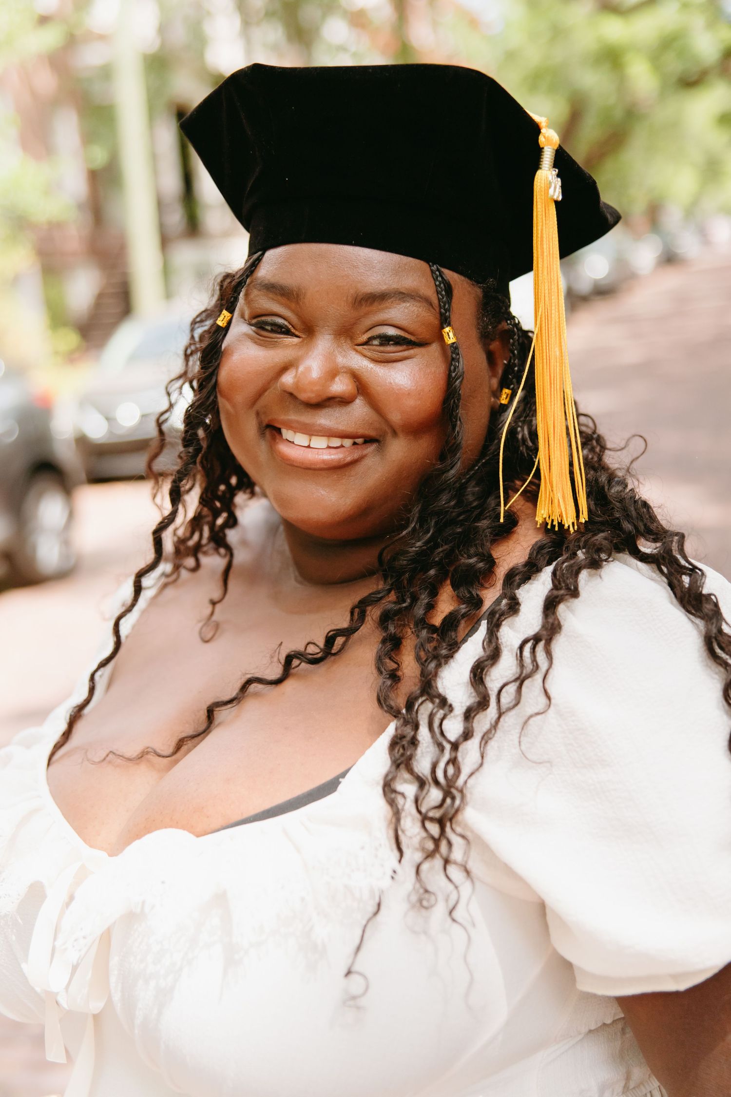 Smiling Black woman wearing black graduation cap with gold tassel in white dress.
