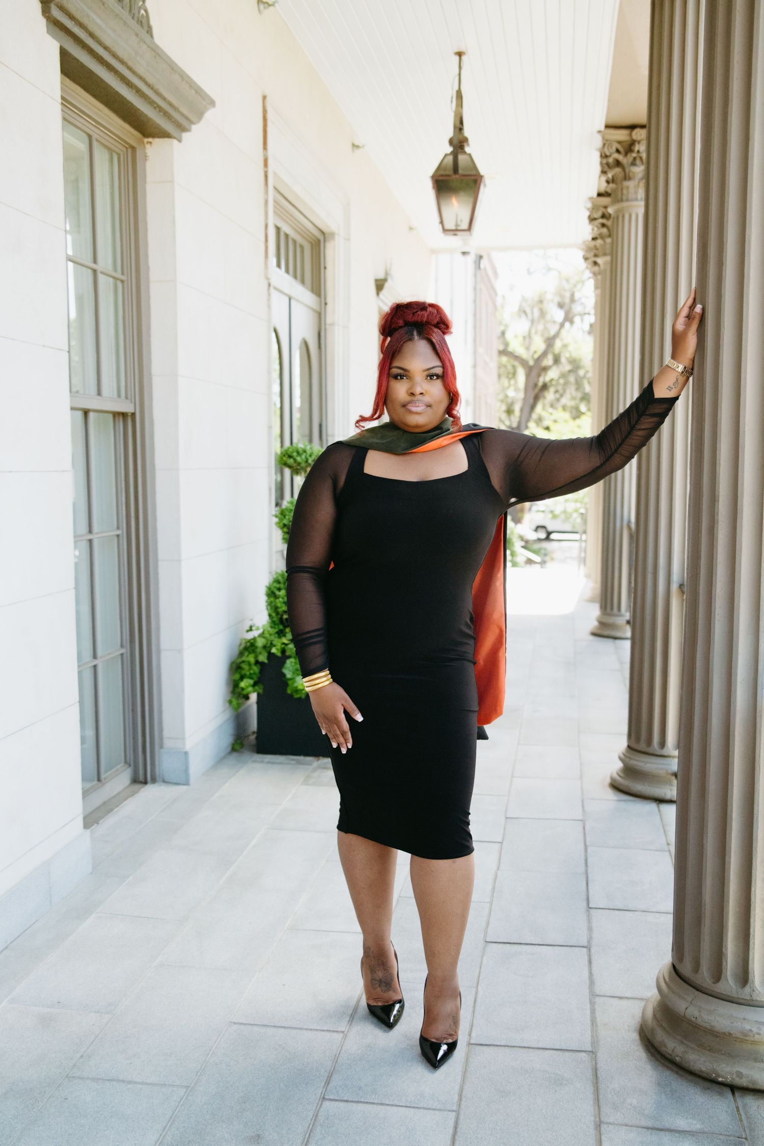 Black woman in black bodycon dress with mesh sleeves posing on building porch.