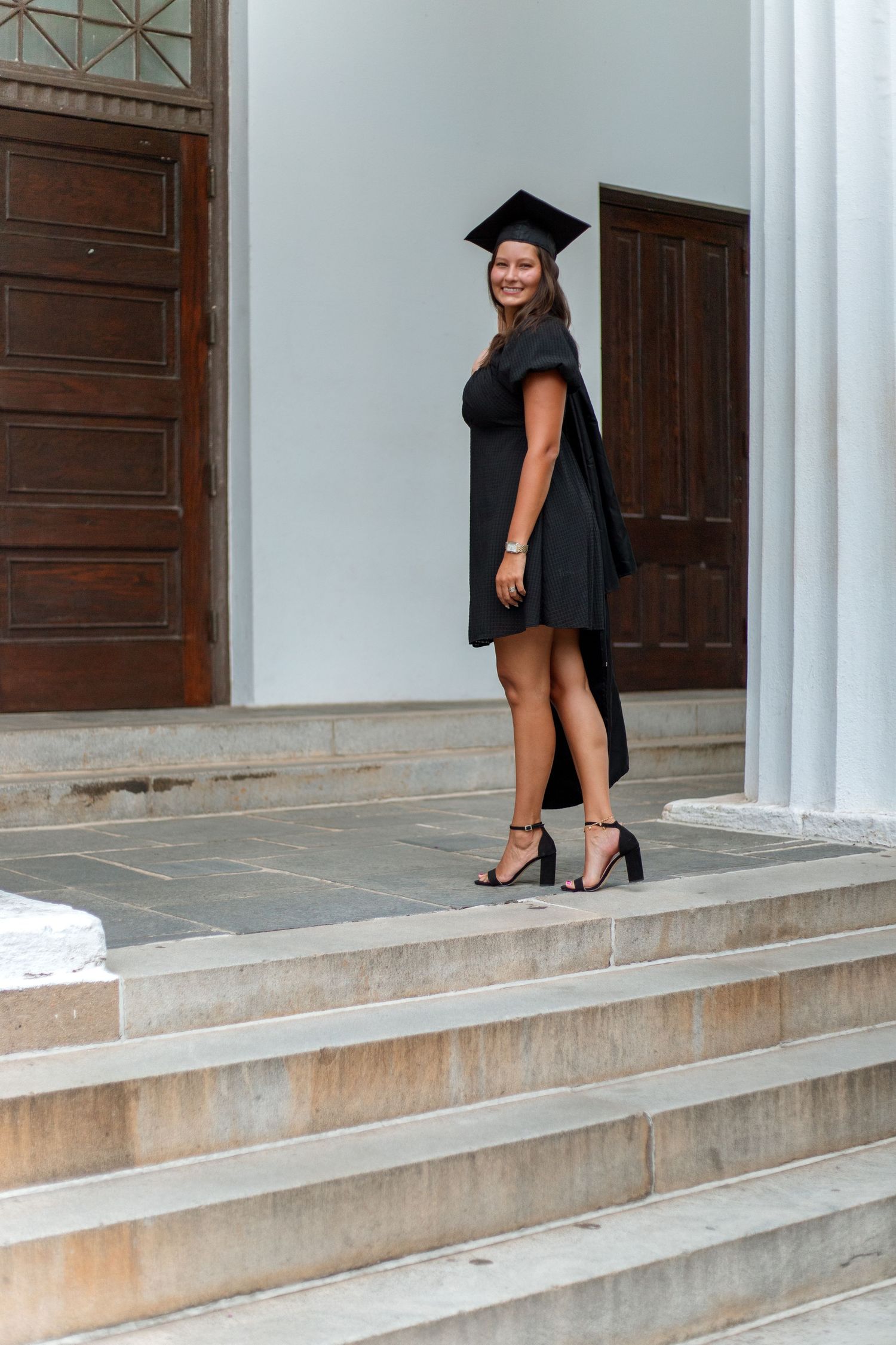 Female graduate in black dress and mortarboard cap posing on steps of white columned building.