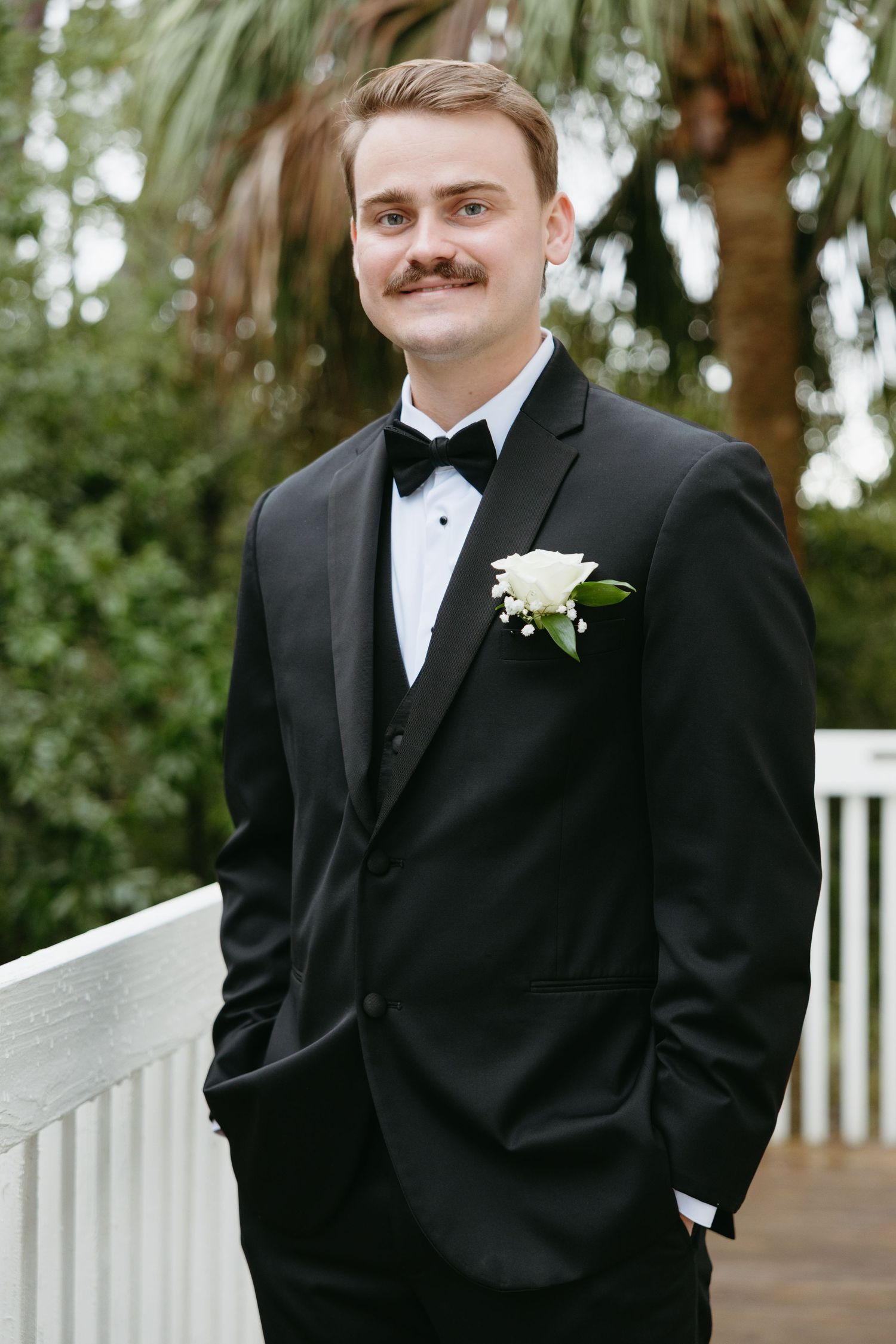 Dapper groom in black tuxedo with white boutonniere smiling outdoors.