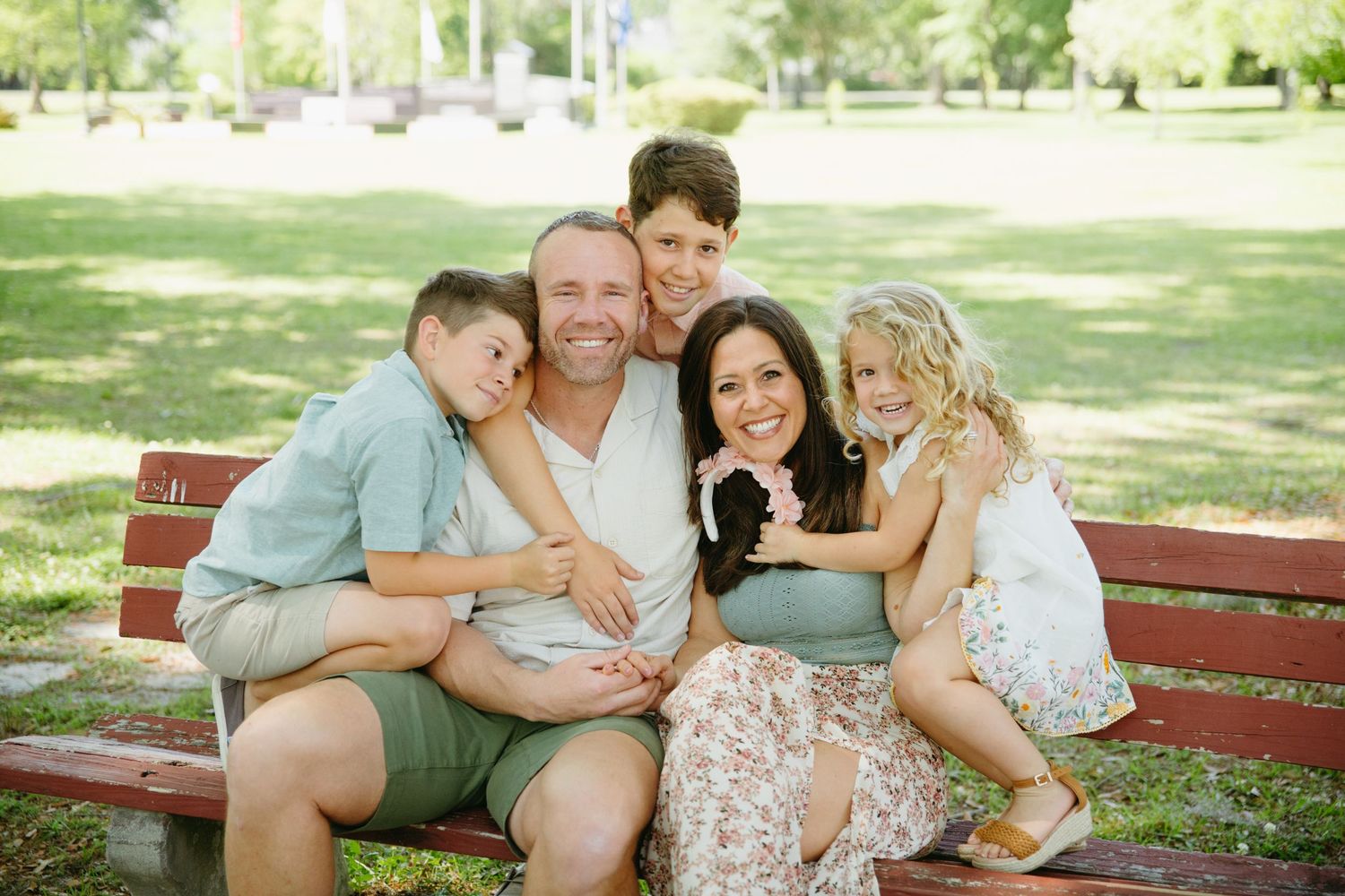Family of five sits together on a red park bench on a sunny green lawn.