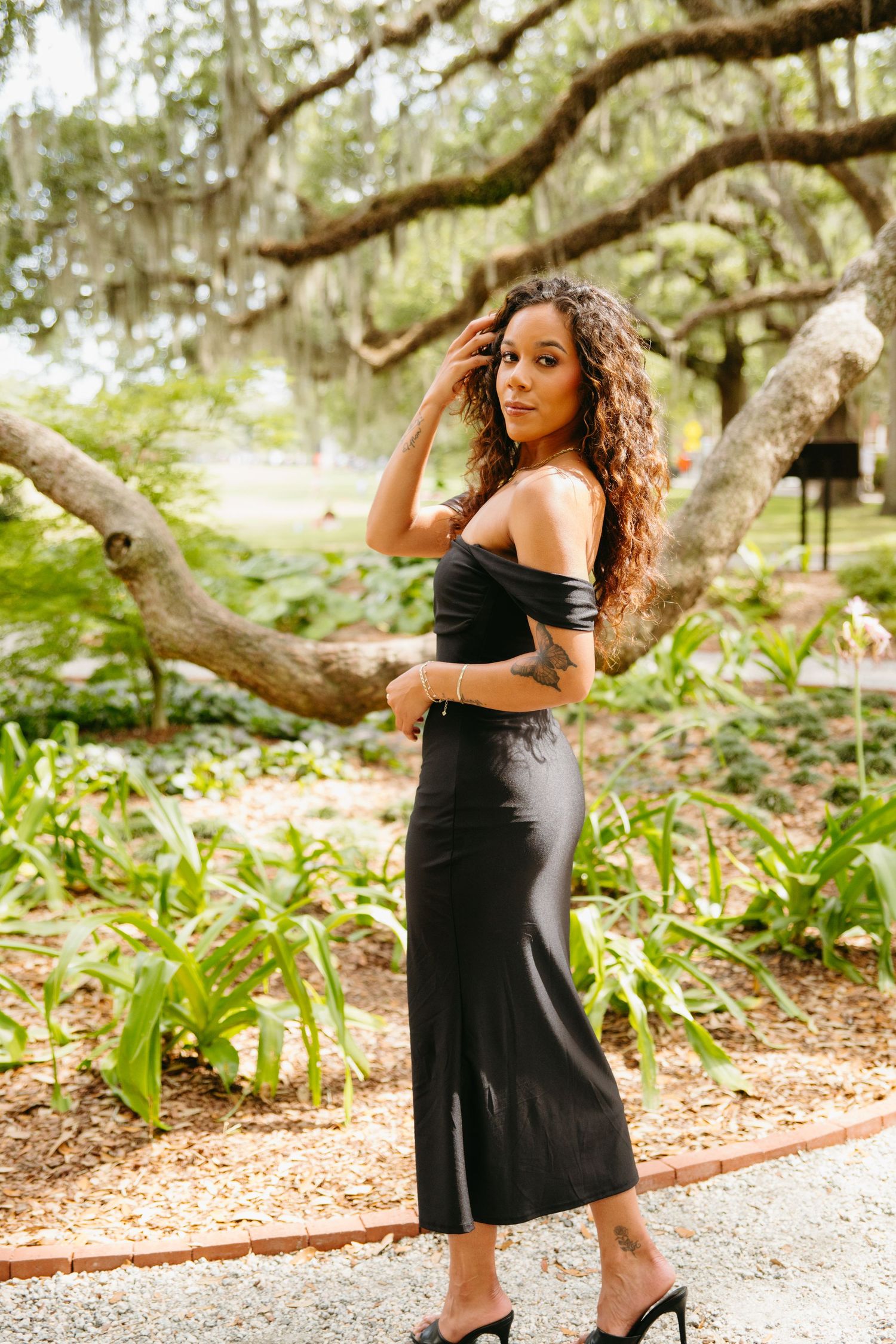 Young woman in black off-shoulder midi dress poses in lush garden setting.