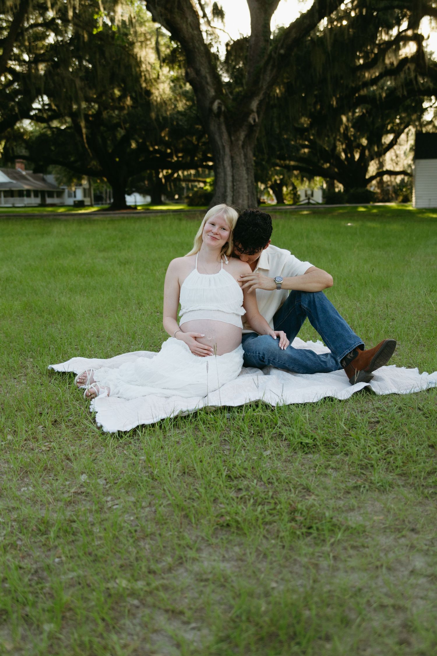 Pregnant couple sitting on a white blanket in a lush green park under large oak trees.