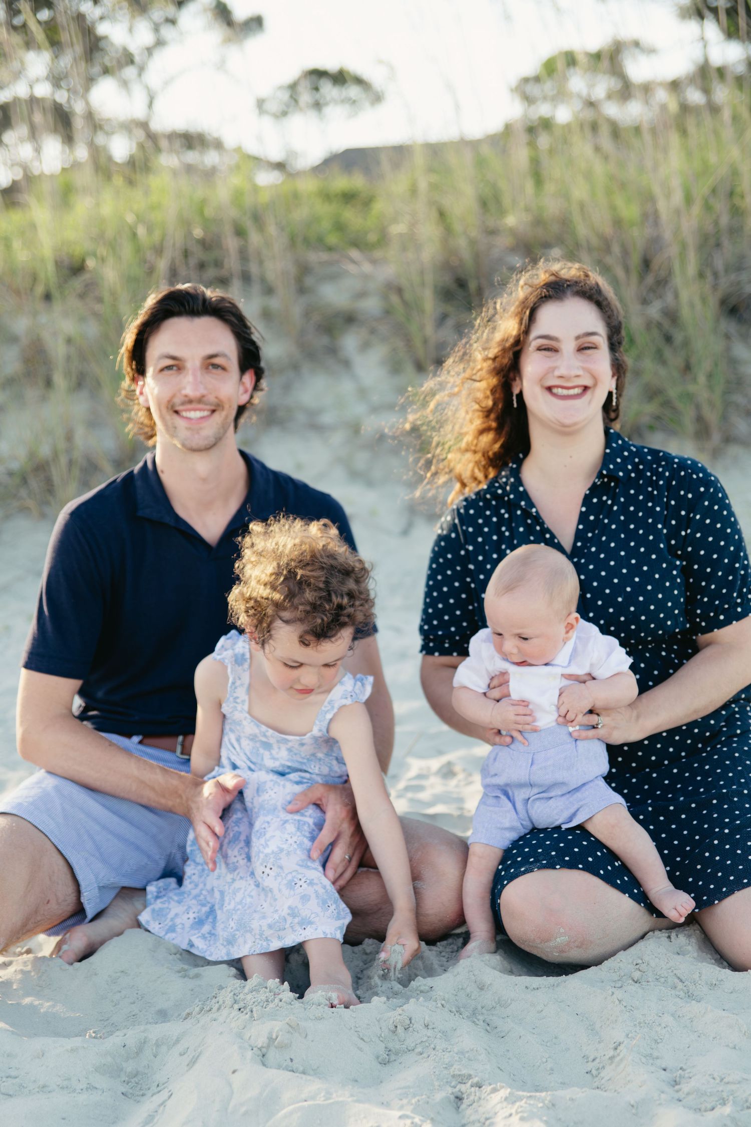 Young family of four sitting on beach sand, parents with toddler girl and baby boy.