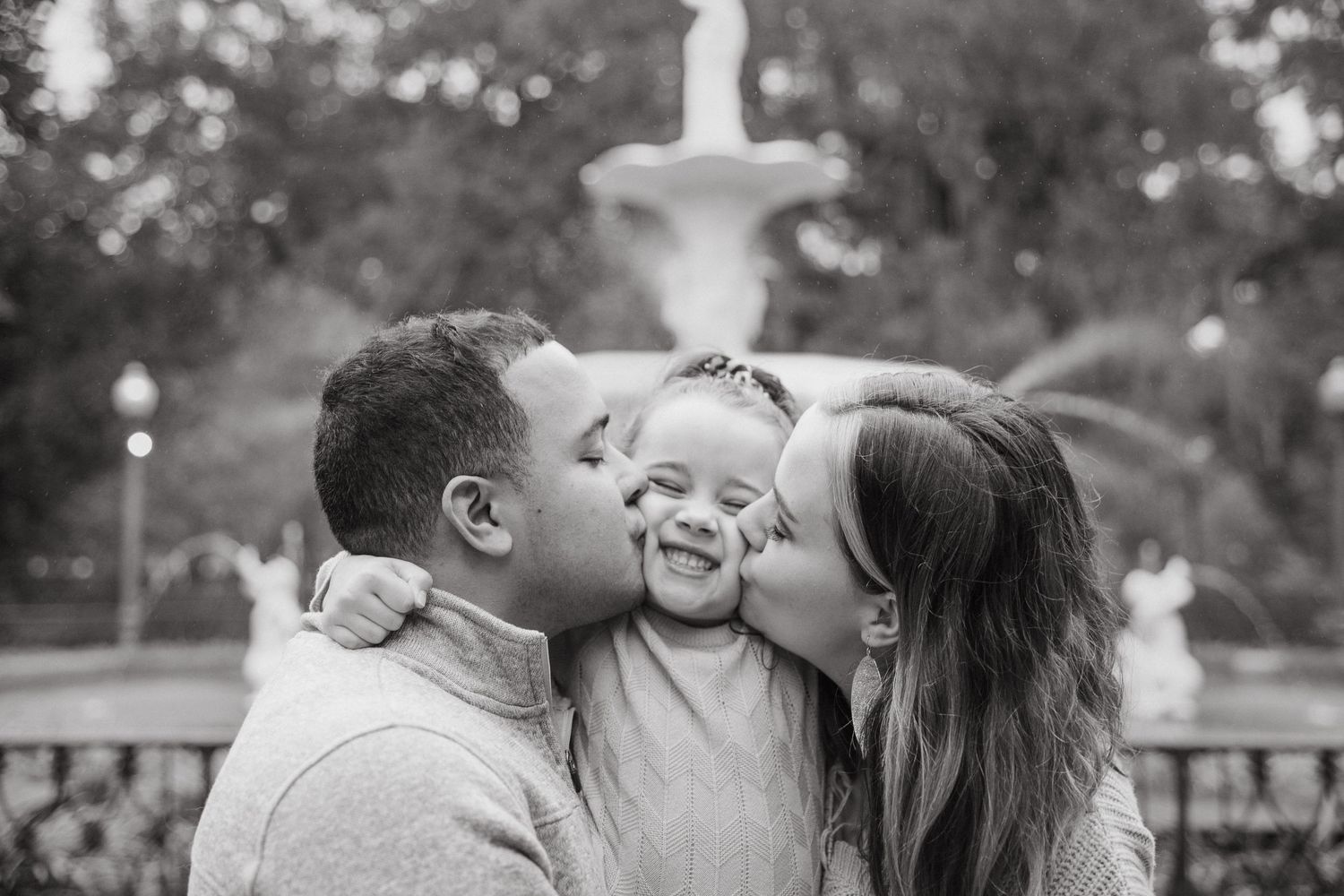 Black and white photo of happy parents kissing their laughing toddler on the cheeks in a park.