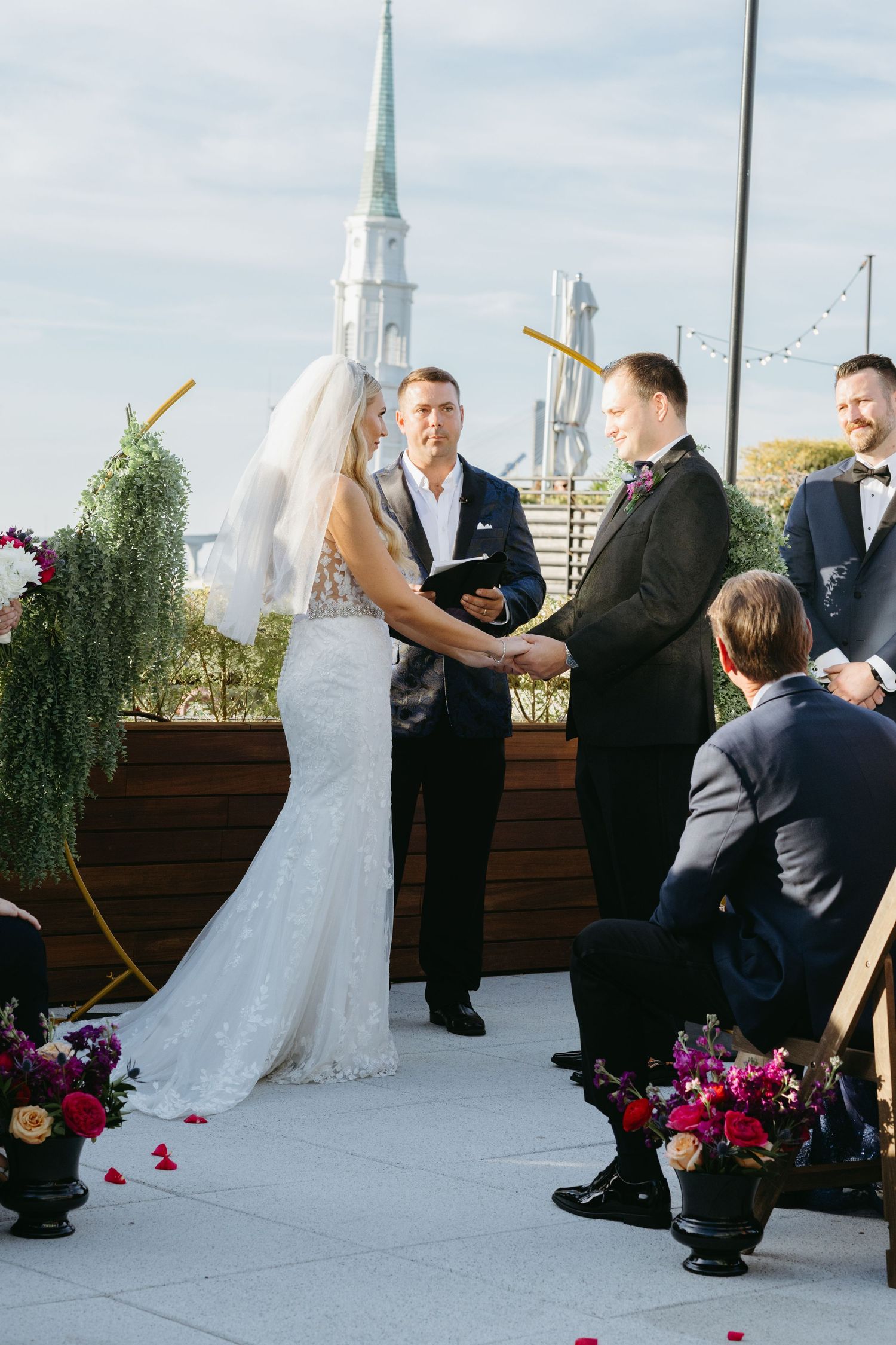 Bride and groom exchanging vows at rooftop ceremony with church steeple in background.