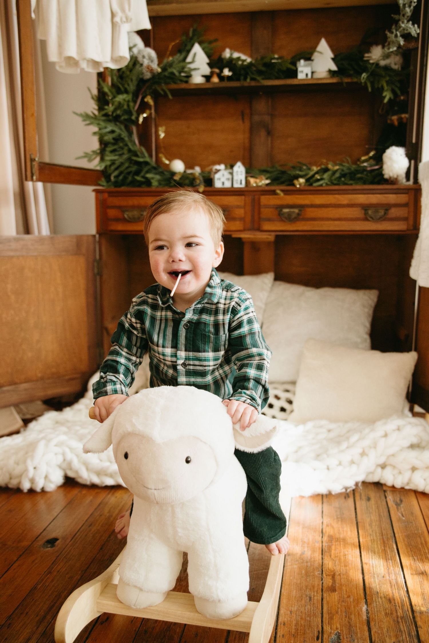 A happy toddler in a green plaid shirt sits on a white lamb rocking toy in a rustic Christmas-decorated room.