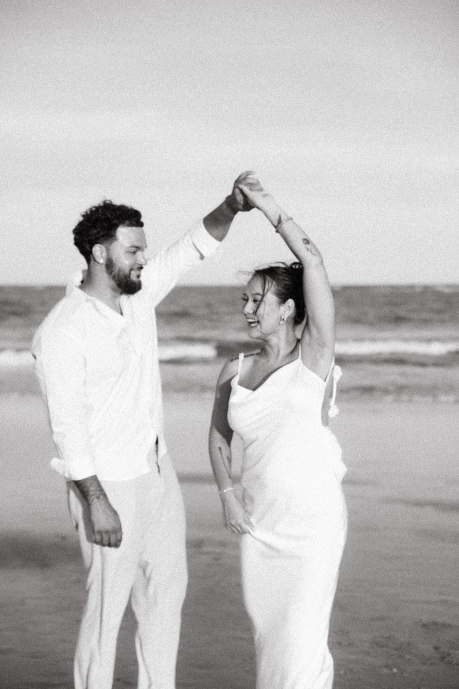 Black and white photo of couple dancing on beach, man spinning woman in white dress.