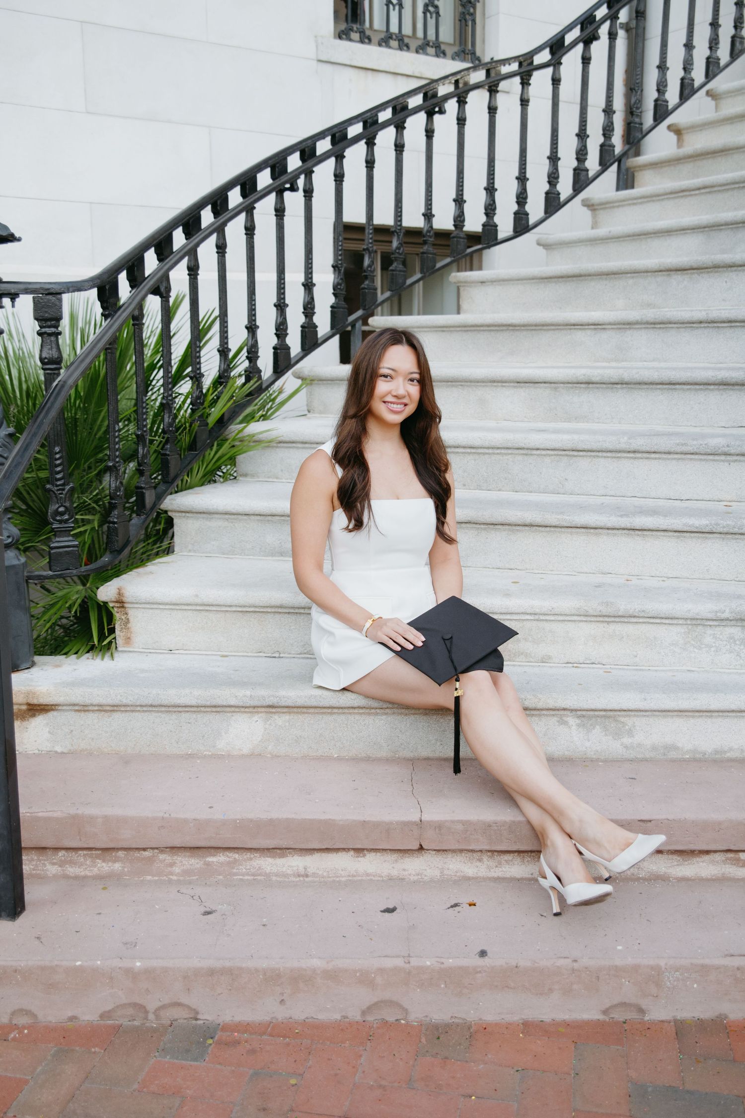Smiling graduate in white dress holding cap while sitting on marble steps.