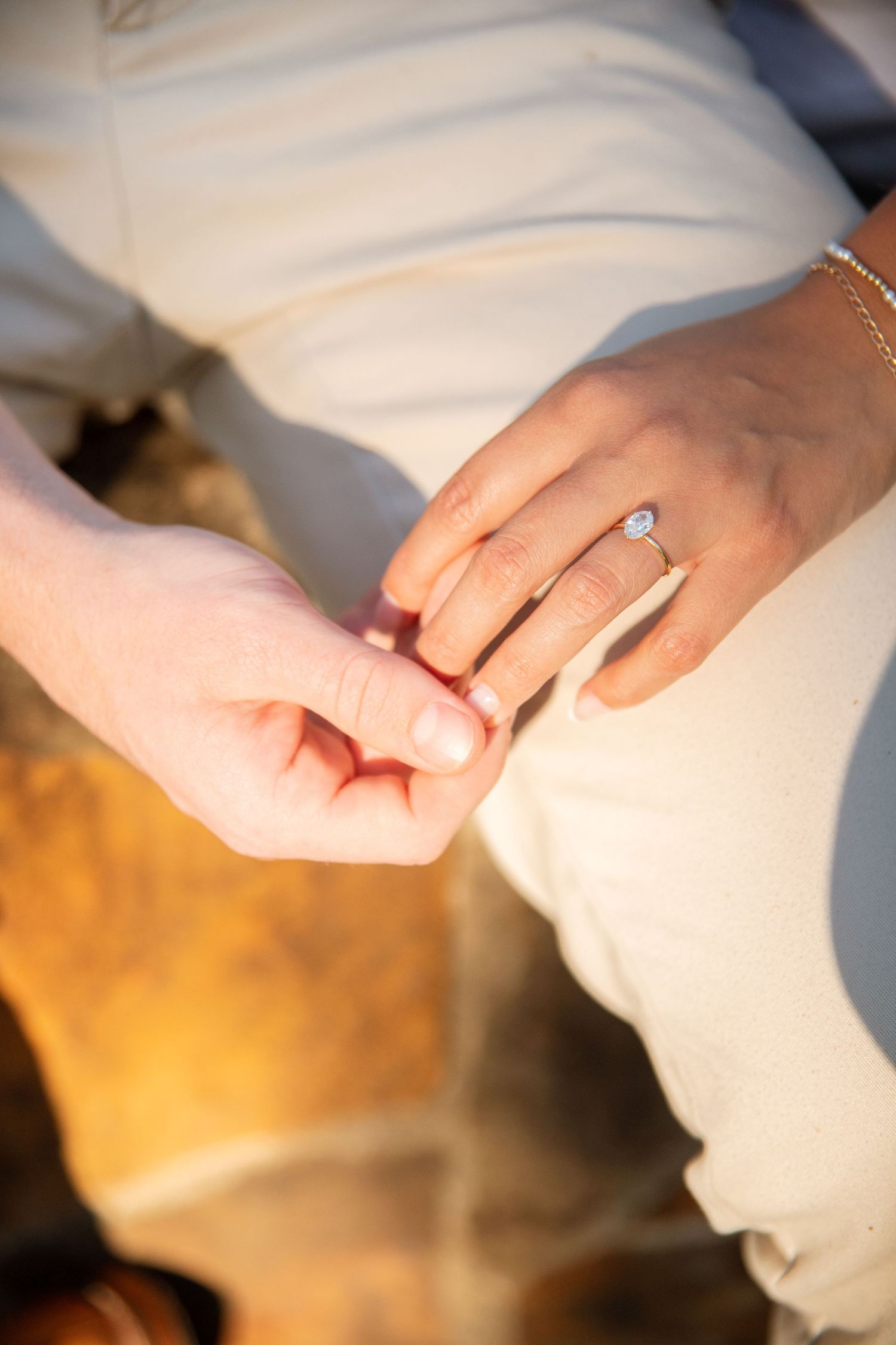 Close-up of couple holding hands showing elegant oval diamond engagement ring in warm golden light.
