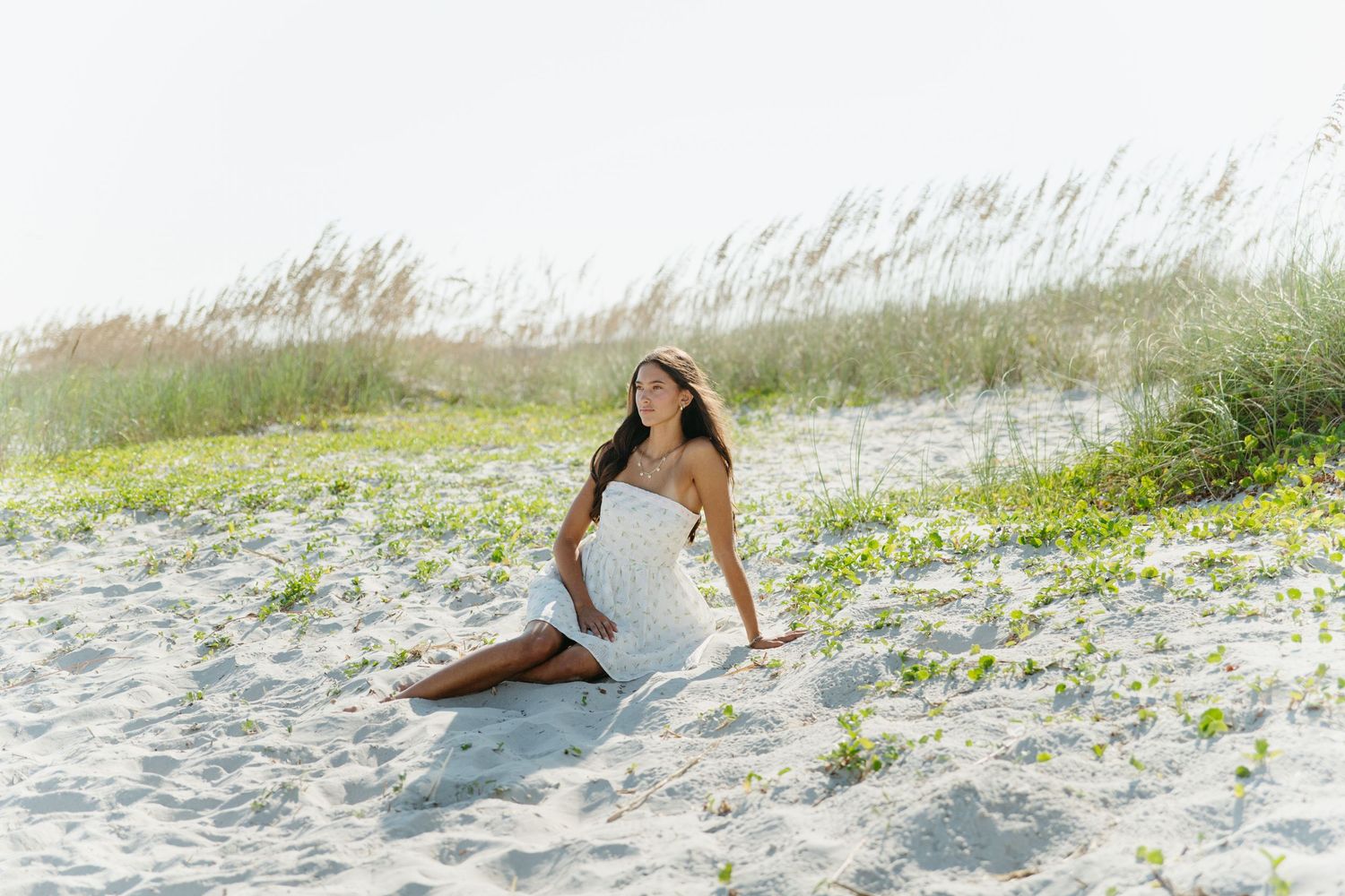 Young woman in a white sundress sitting gracefully on sandy beach dunes with sea oats behind her.