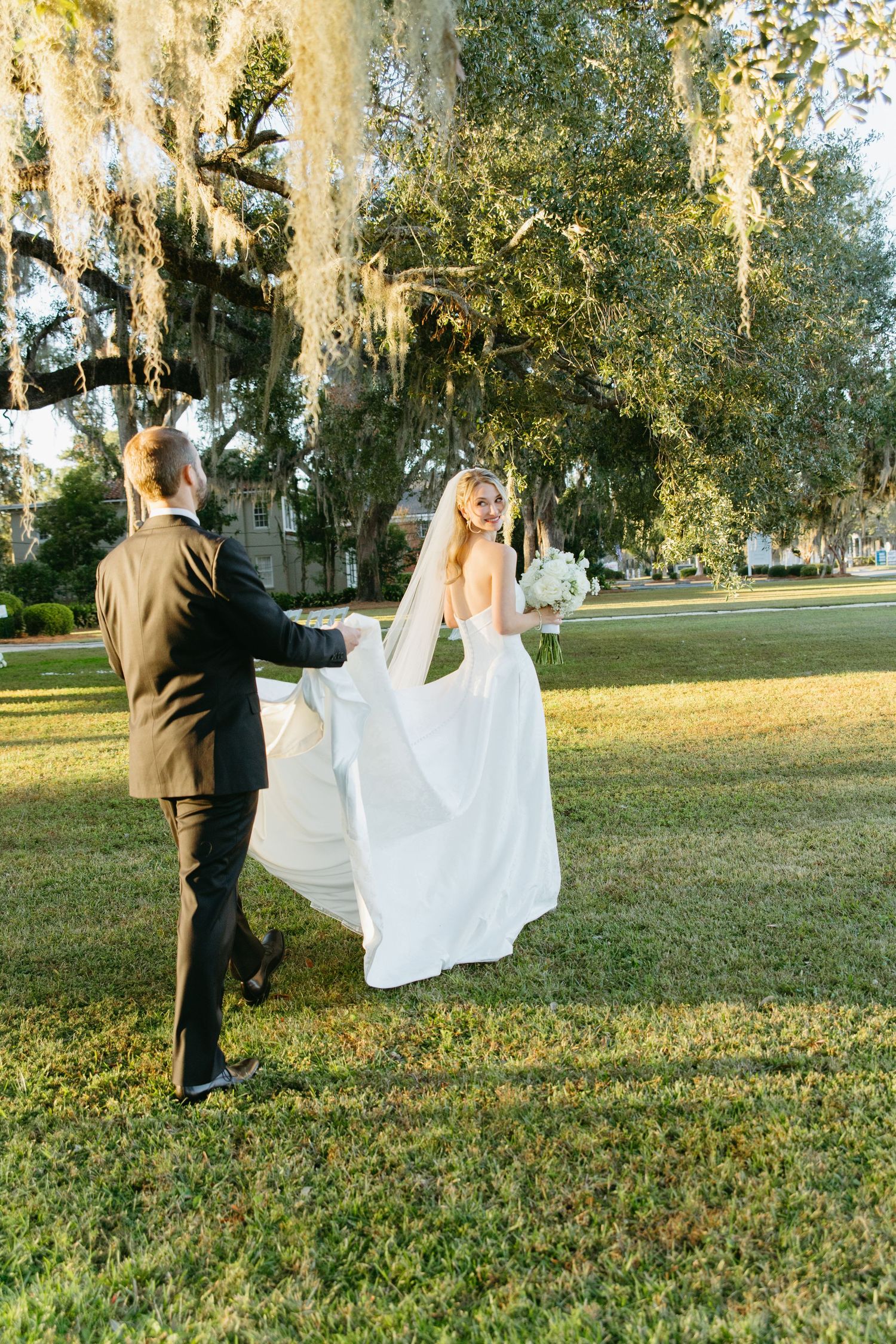 Groom helping bride with her flowing white gown under Spanish moss-draped oak trees on green lawn.