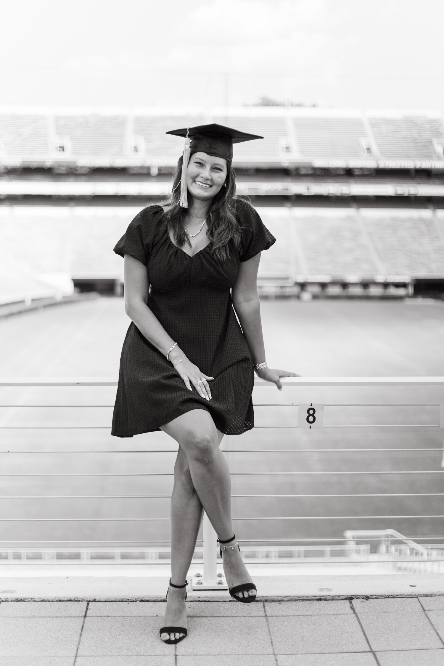 Black and white photo of smiling female graduate in black dress and cap posing at stadium.