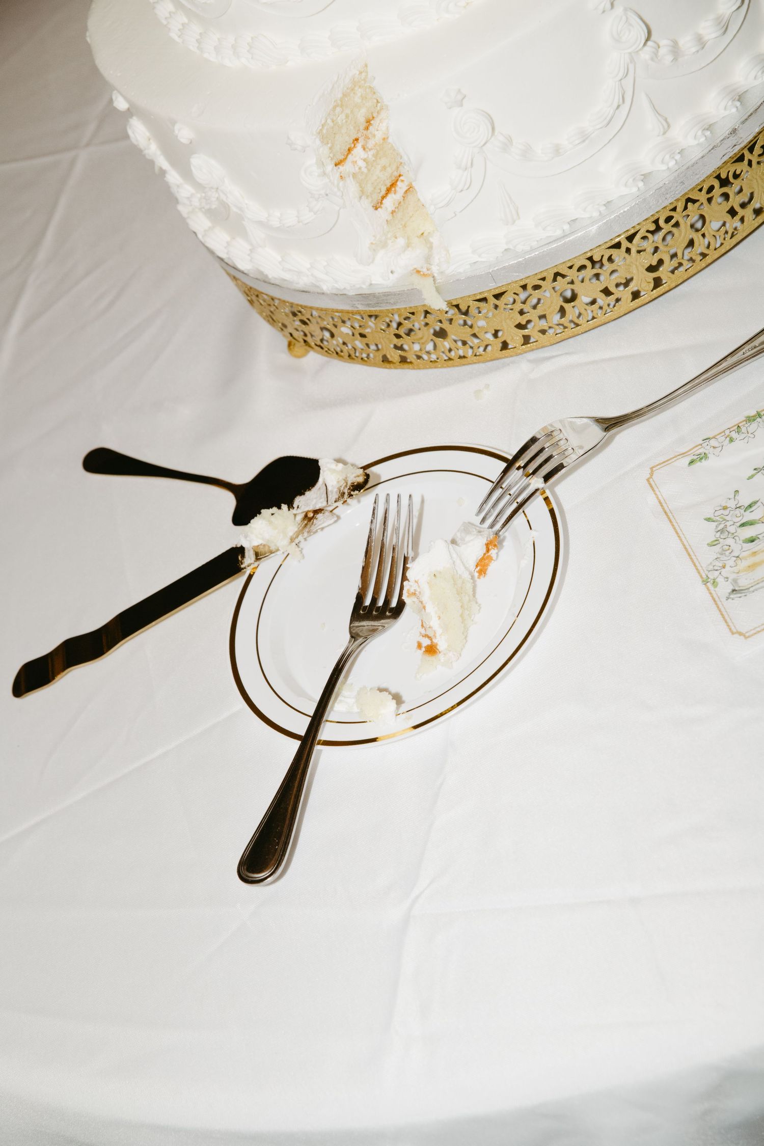 Partially sliced white wedding cake on gold stand with two forks and cake server on elegant plate.