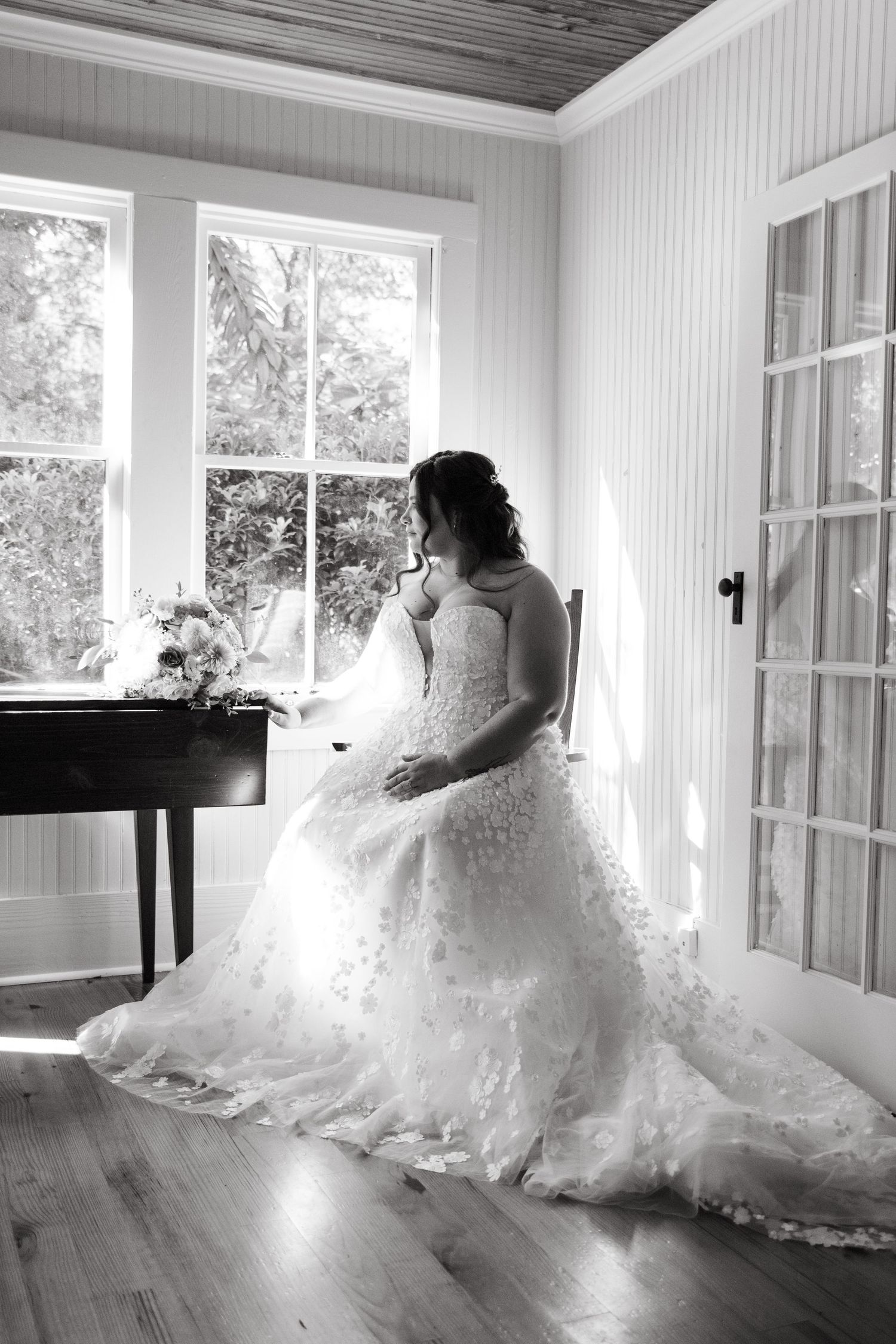 Black and white photo of bride in lace ball gown gazing out sunlit window beside a grand piano.