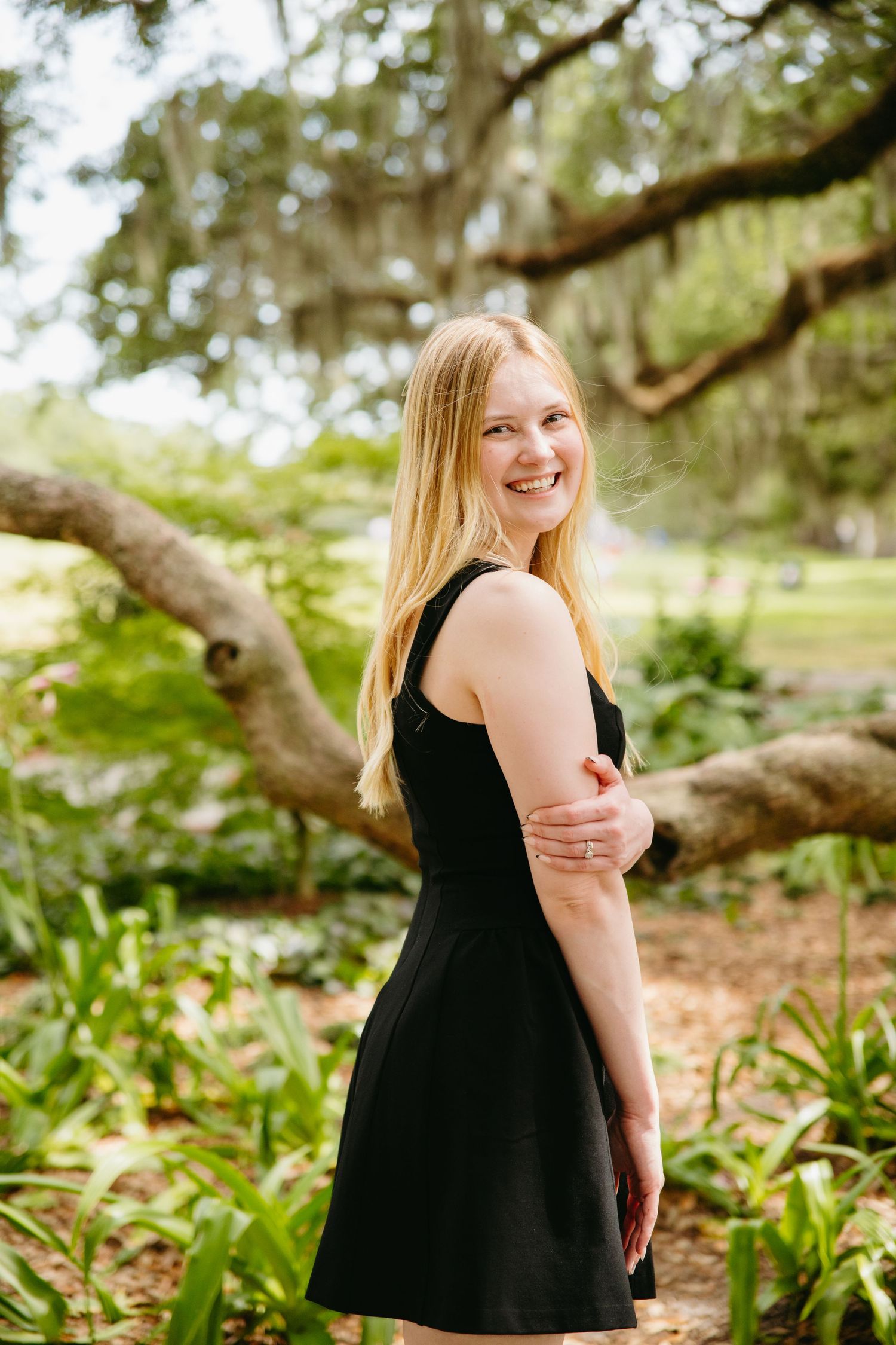 Smiling blonde woman in black dress poses outdoors under moss-covered oak tree.