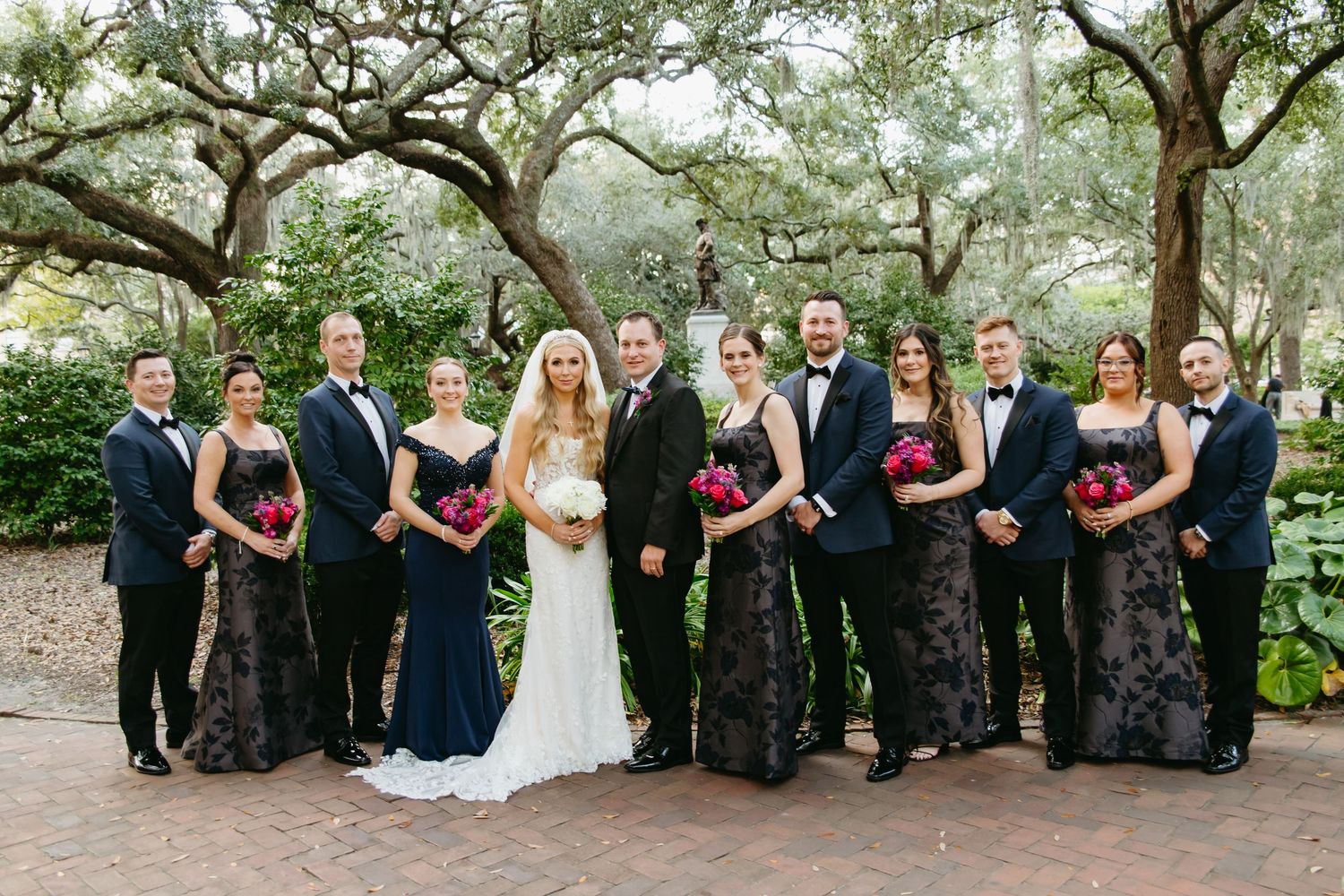 Wedding party in navy and black attire posing under oak trees with vibrant pink bouquets.
