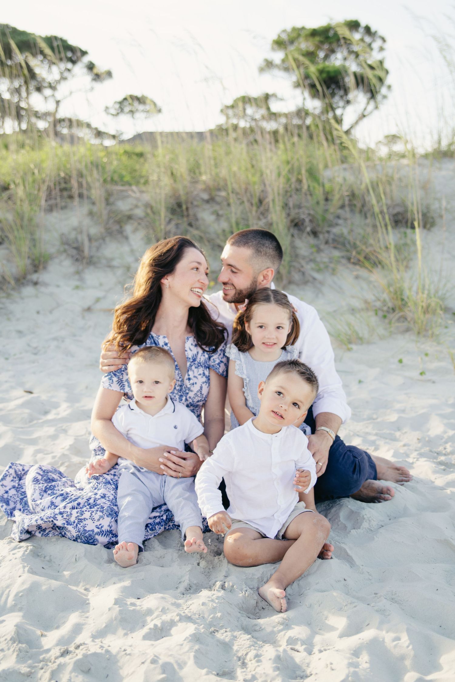 Family of five sitting on beach sand, parents gazing at each other with three young children.