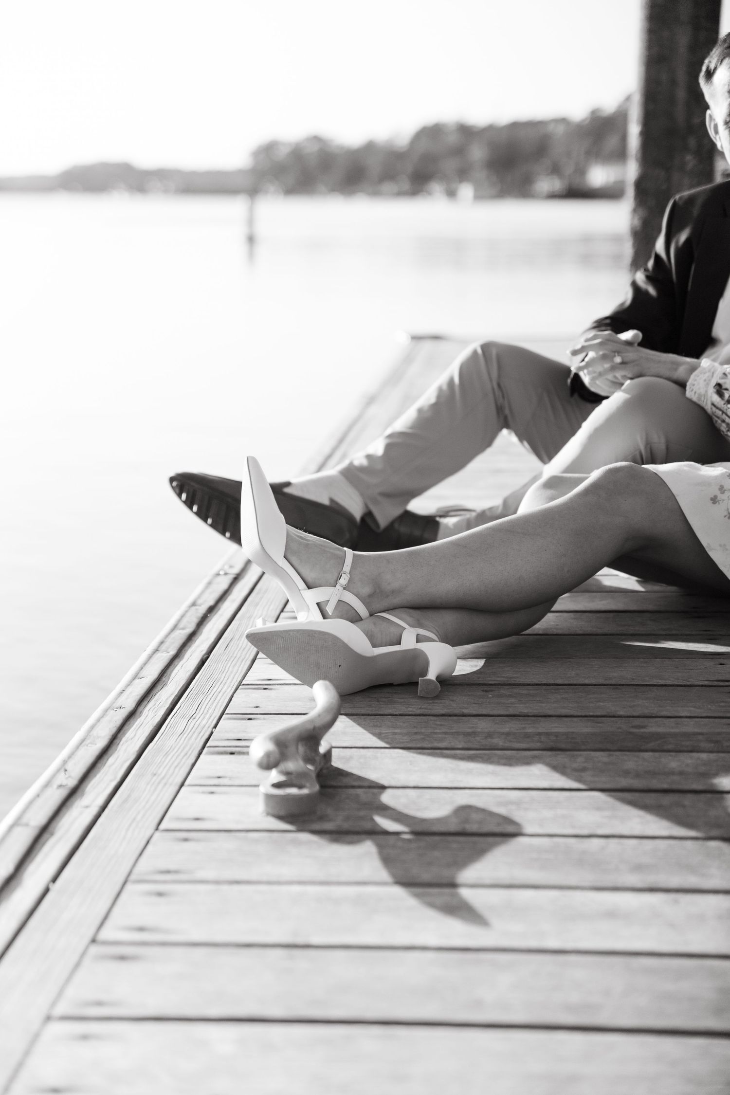 Black and white photo of couple sitting on a dock by the water, shoes removed, legs stretched out.