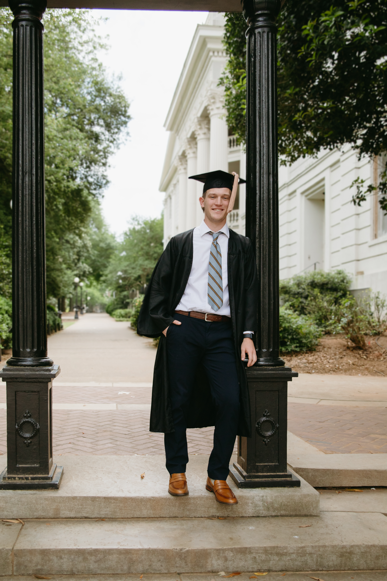 Male graduate in cap and gown leaning against column on tree-lined university campus walkway.