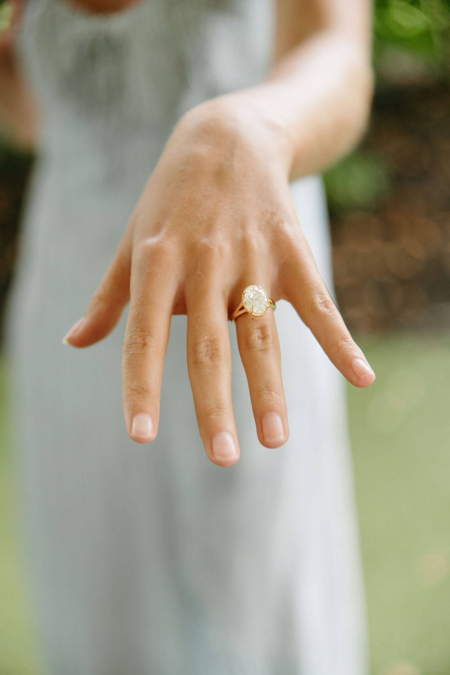 Close-up of a woman\u2019s hand displaying a large oval diamond engagement ring in a gold setting outdoors.