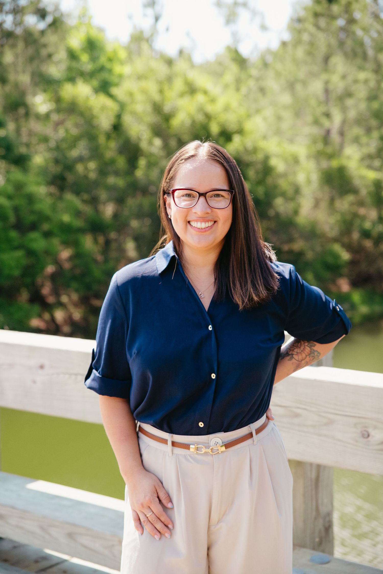 Smiling woman in navy blue shirt and glasses posing on wooden bridge with green nature background.