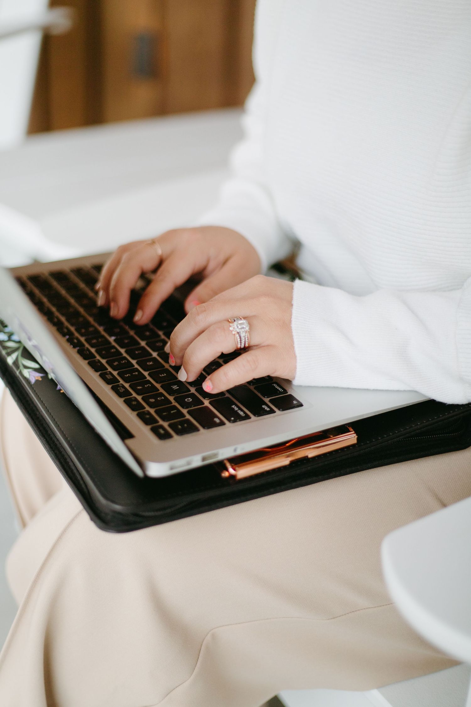 Close-up of hands with diamond engagement ring typing on a rose gold laptop in a black leather folio.
