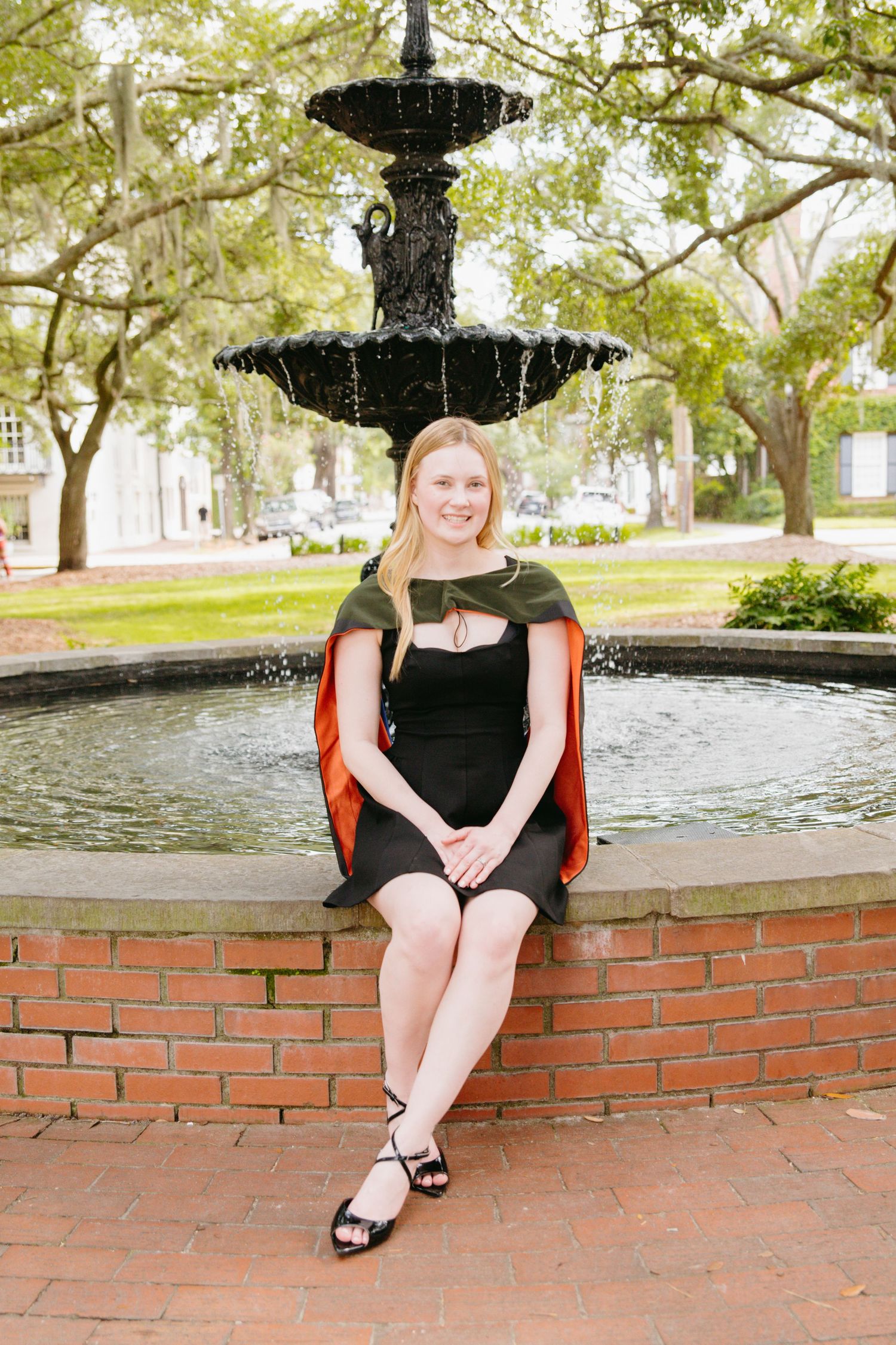 Blonde female graduate in black dress and academic hood sits by ornate fountain.