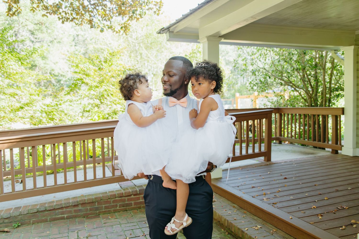 Groom in black suit holds two young girls in white tutus on a wooden deck surrounded by greenery.