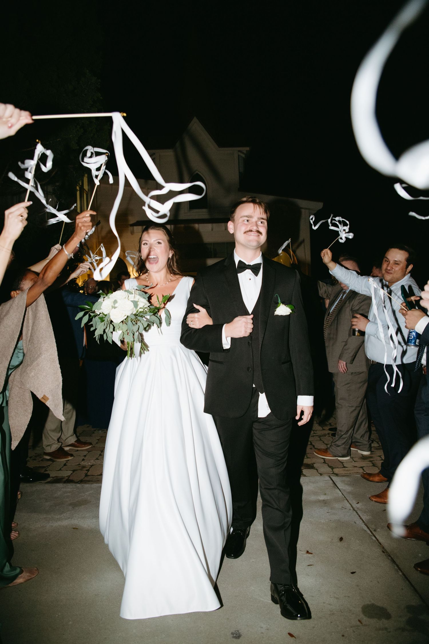 Bride and groom exit wedding ceremony through ribbon wand send-off tunnel at night.