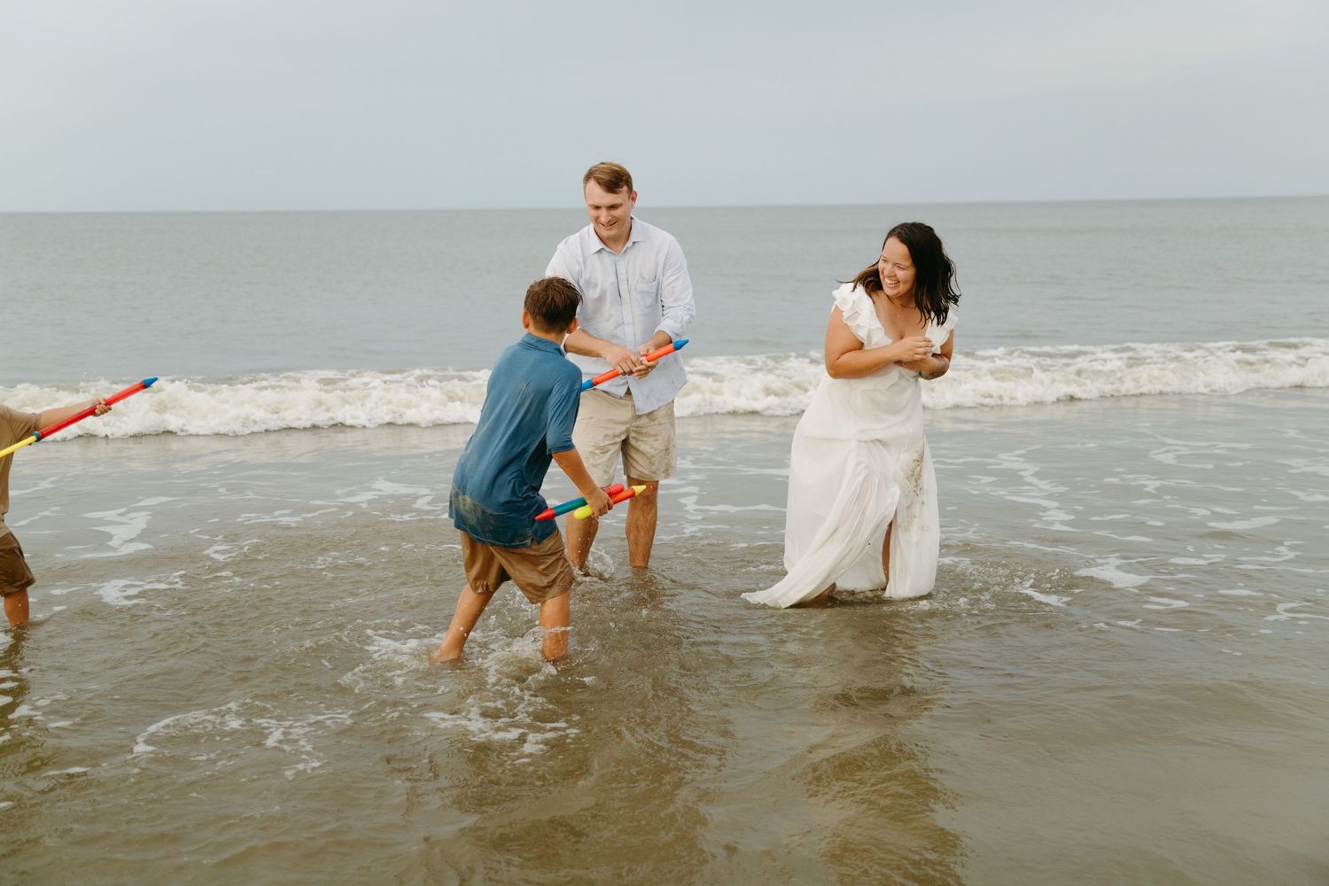 Family enjoys playful moment in shallow ocean waves on a cloudy beach day.