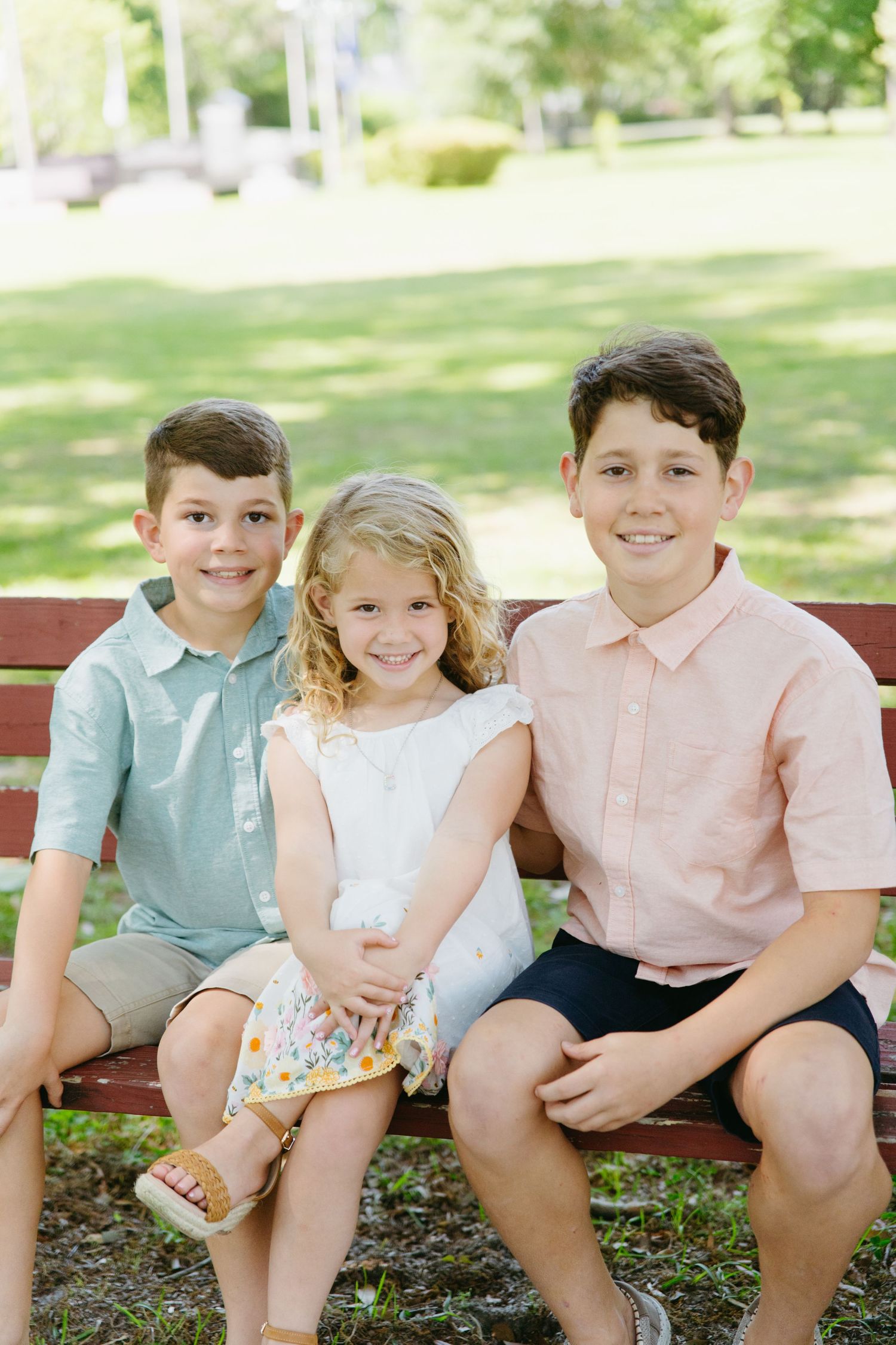 Three siblings, two boys and a girl, smiling together on a park bench outdoors.