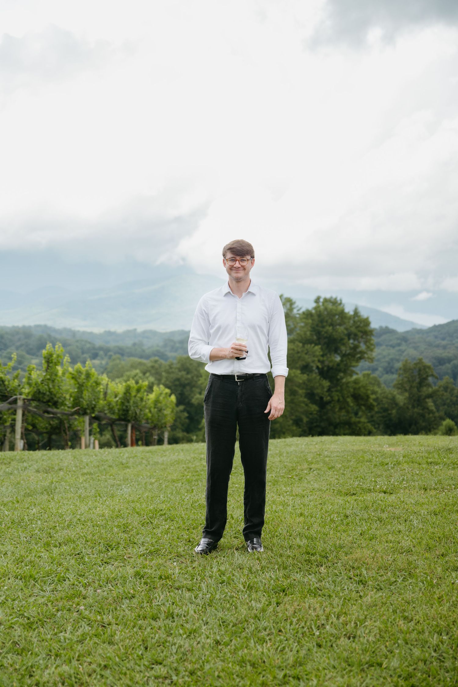Young man in white dress shirt and black pants stands smiling on a green lawn with mountain views behind him.