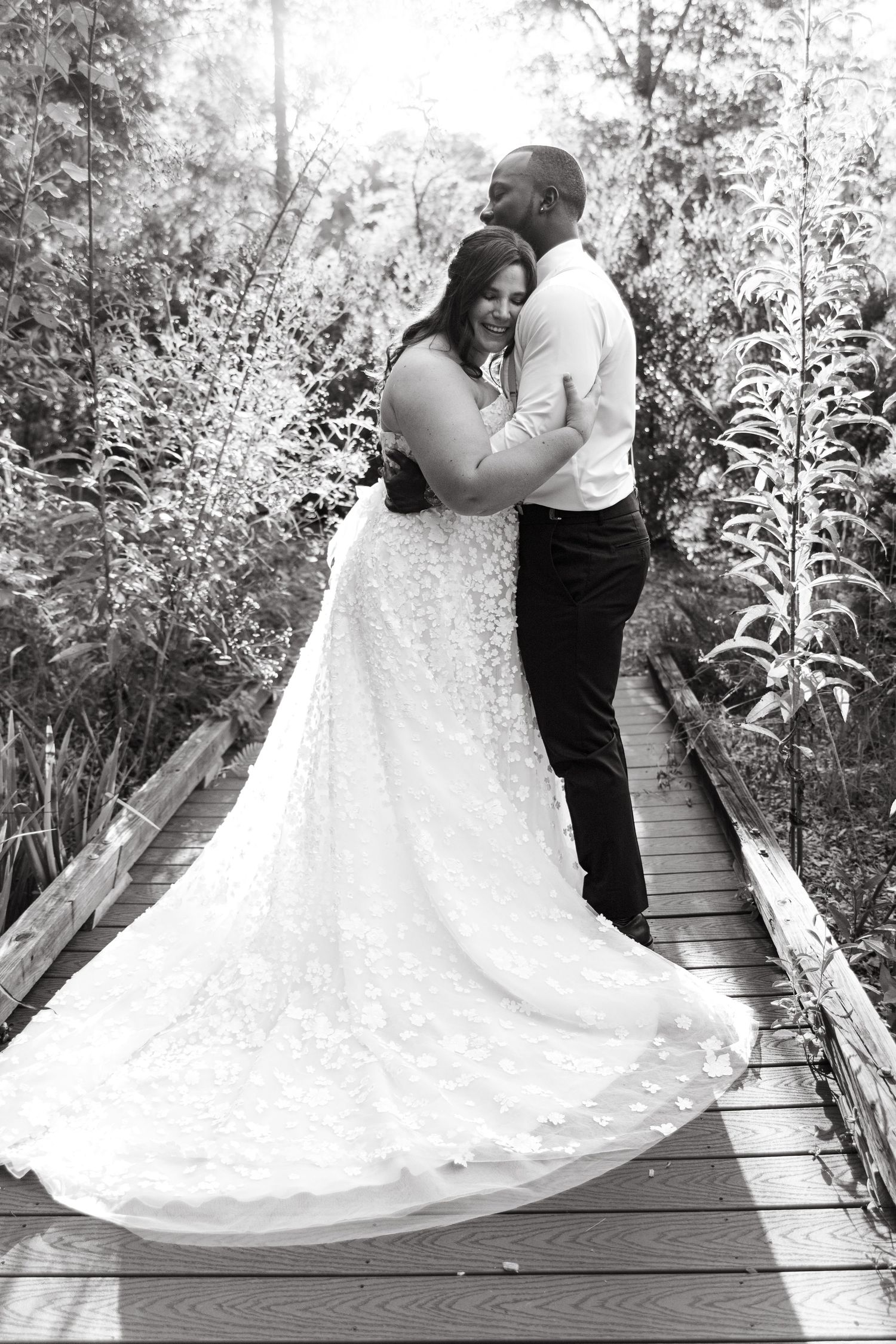 Couple embracing on a wooden bridge surrounded by lush greenery in a romantic black and white photo.