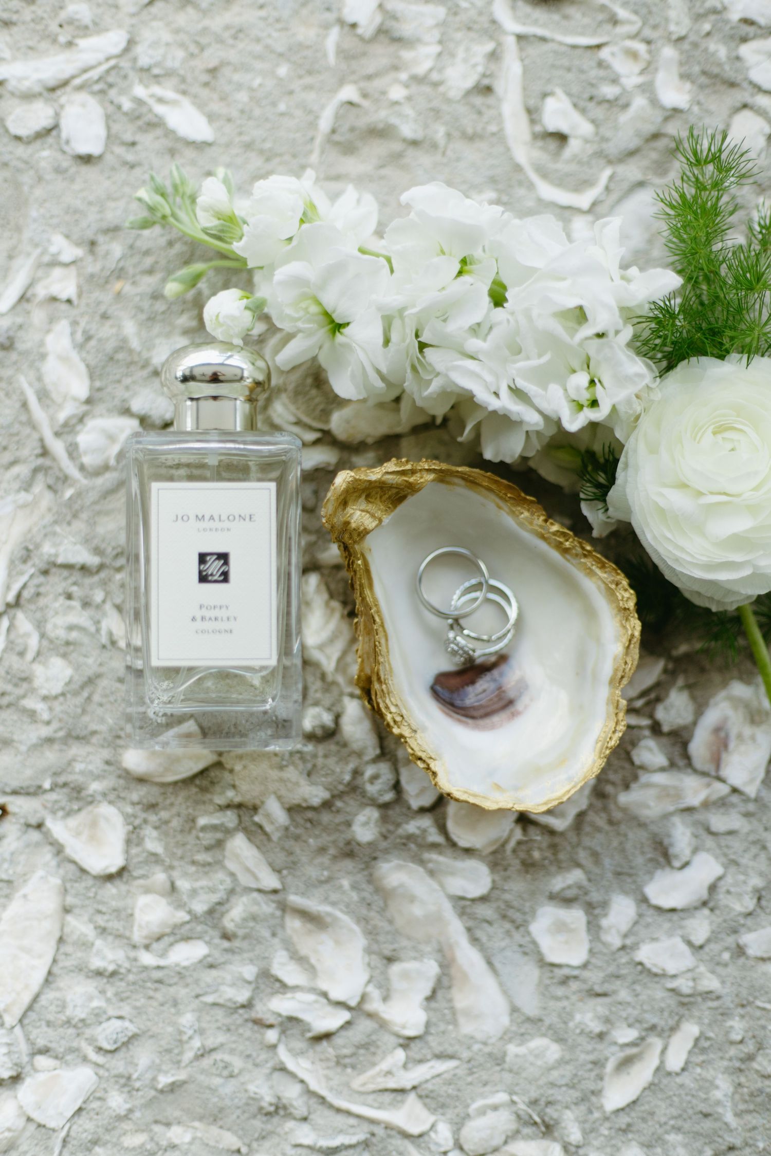 Wedding flat lay with Jo Malone perfume, rings in gold oyster shell, and white flowers on stone surface.