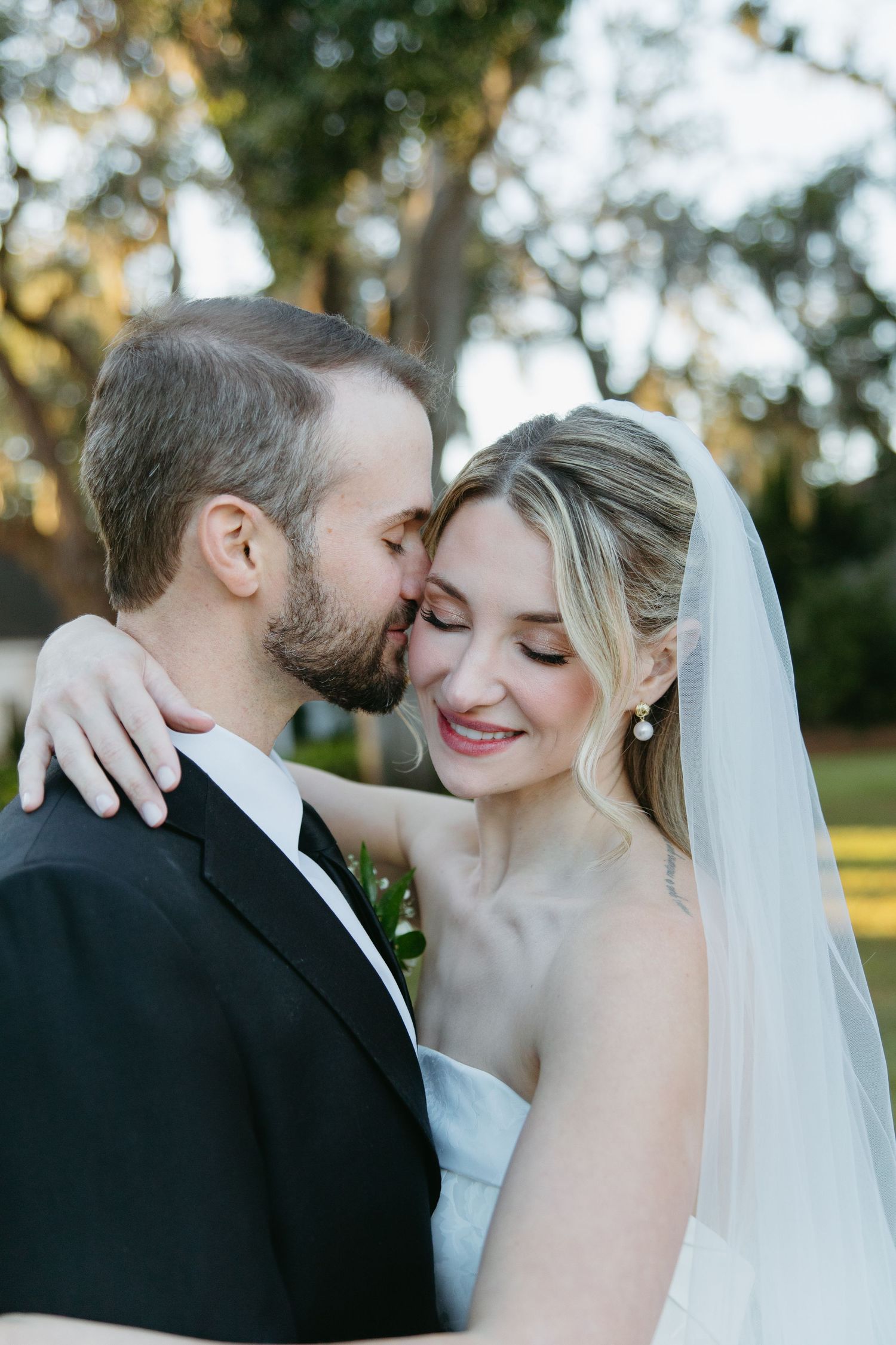 Groom kisses bride on forehead as she smiles with eyes closed wearing a white veil and strapless gown outdoors.