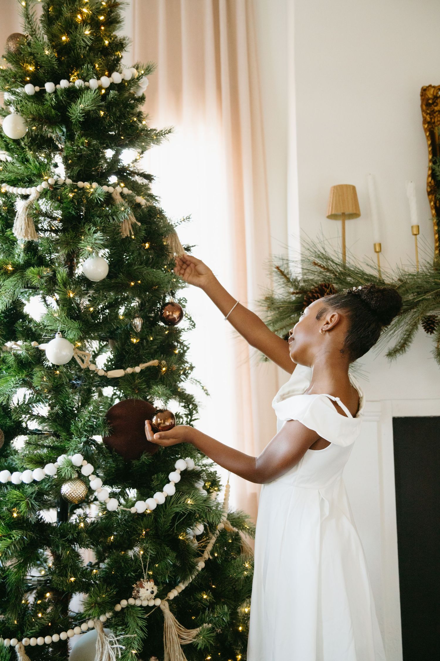 A young girl in a white dress reaches up to place ornaments on a beautifully decorated Christmas tree with pearl garland.