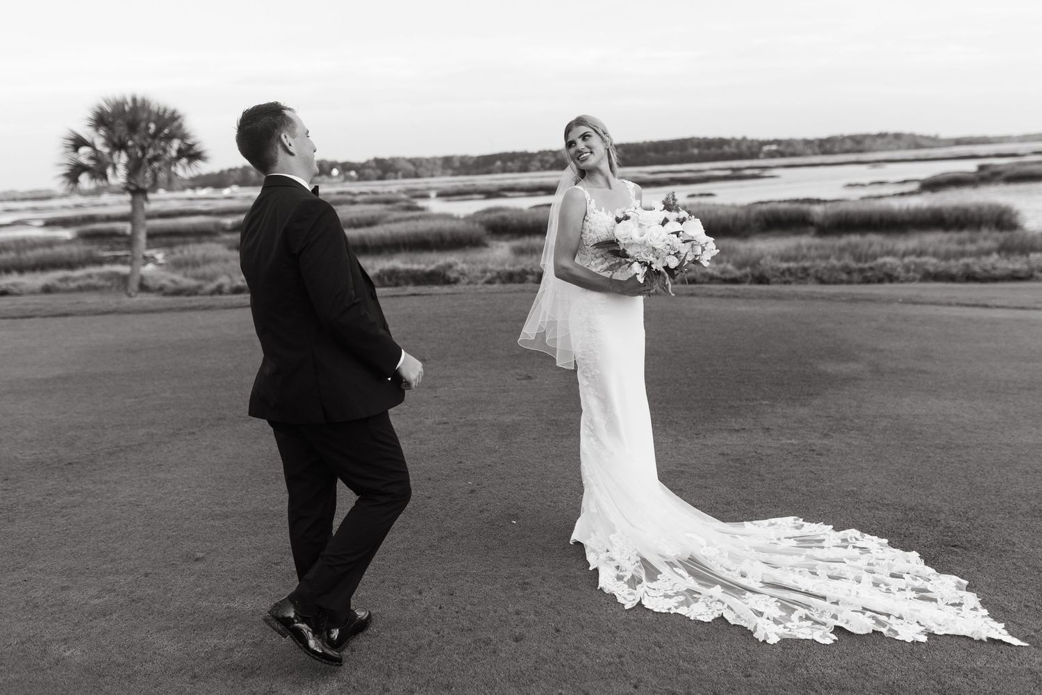 Black and white photo of a bride in a lace gown holding bouquet during first look with groom outdoors.