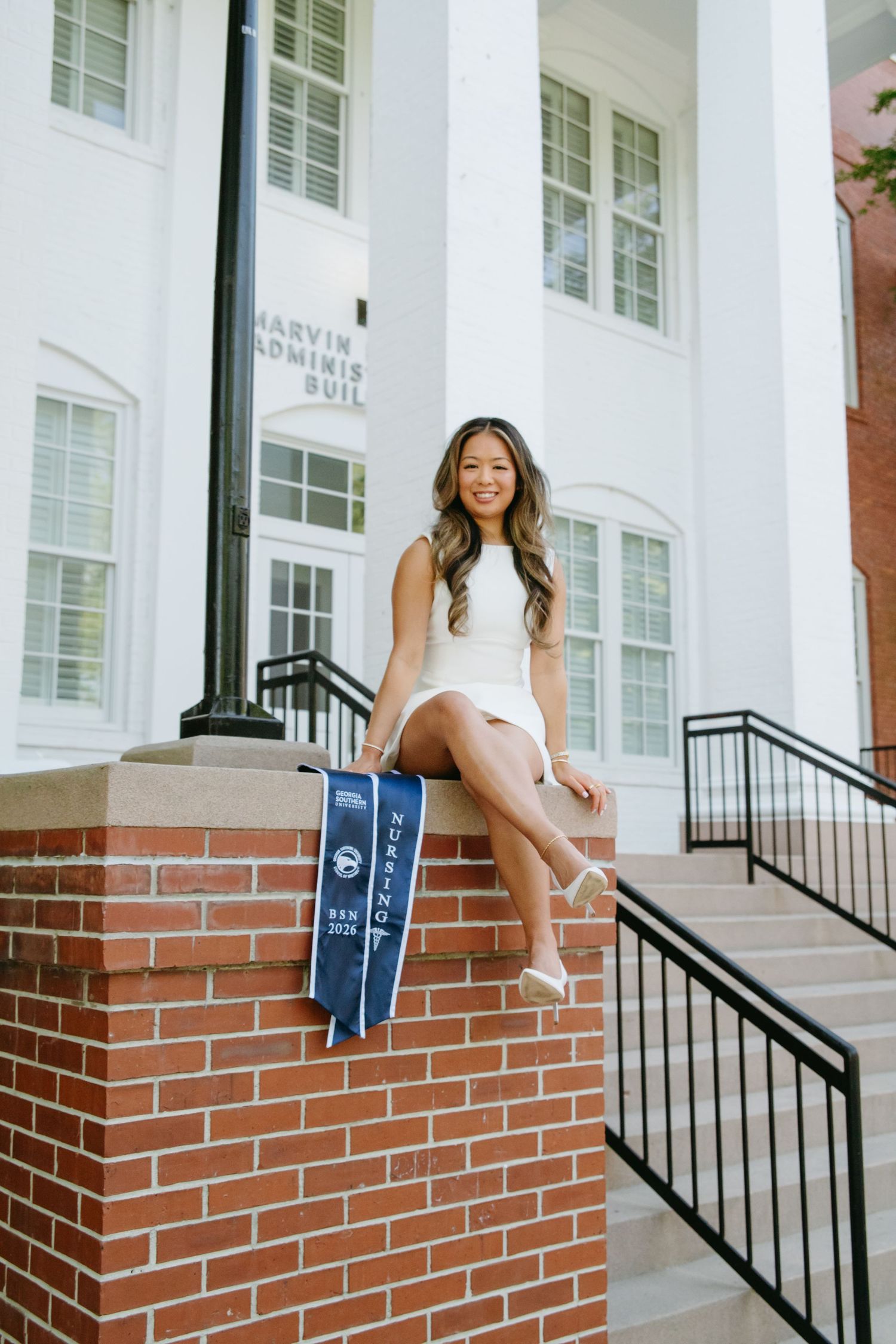 Smiling graduate in white dress sitting on brick wall with blue graduation stole outside Marvin Administration Building.