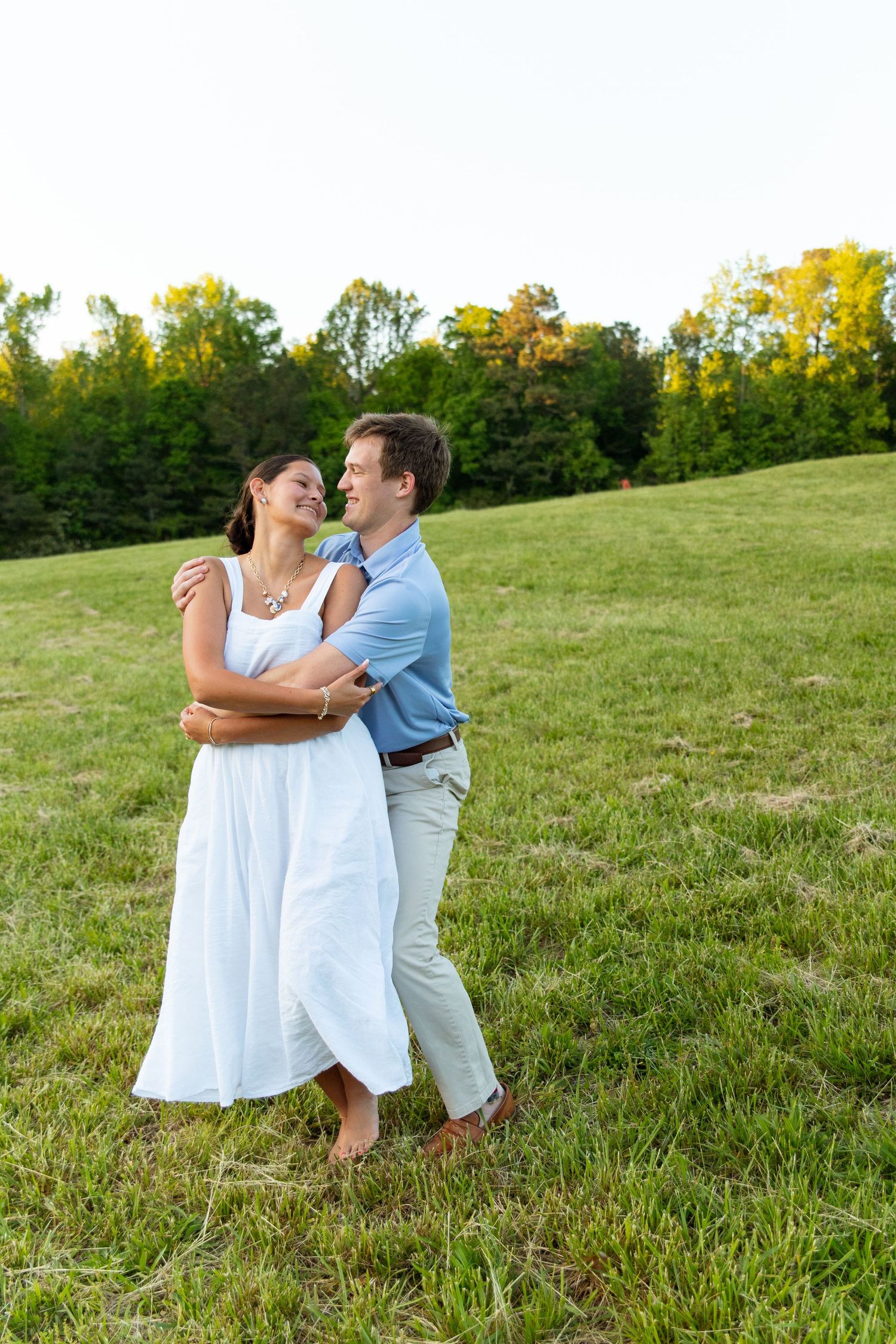 Happy couple embracing and laughing in a green meadow with trees in the background at sunset.