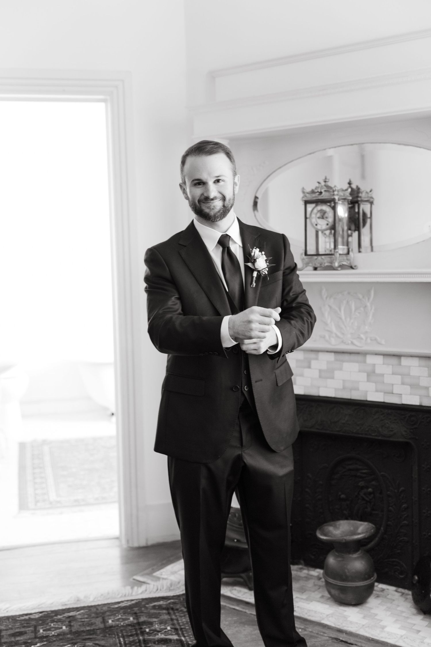 Smiling groom in black tuxedo with boutonniere stands by ornate fireplace in black and white photo.