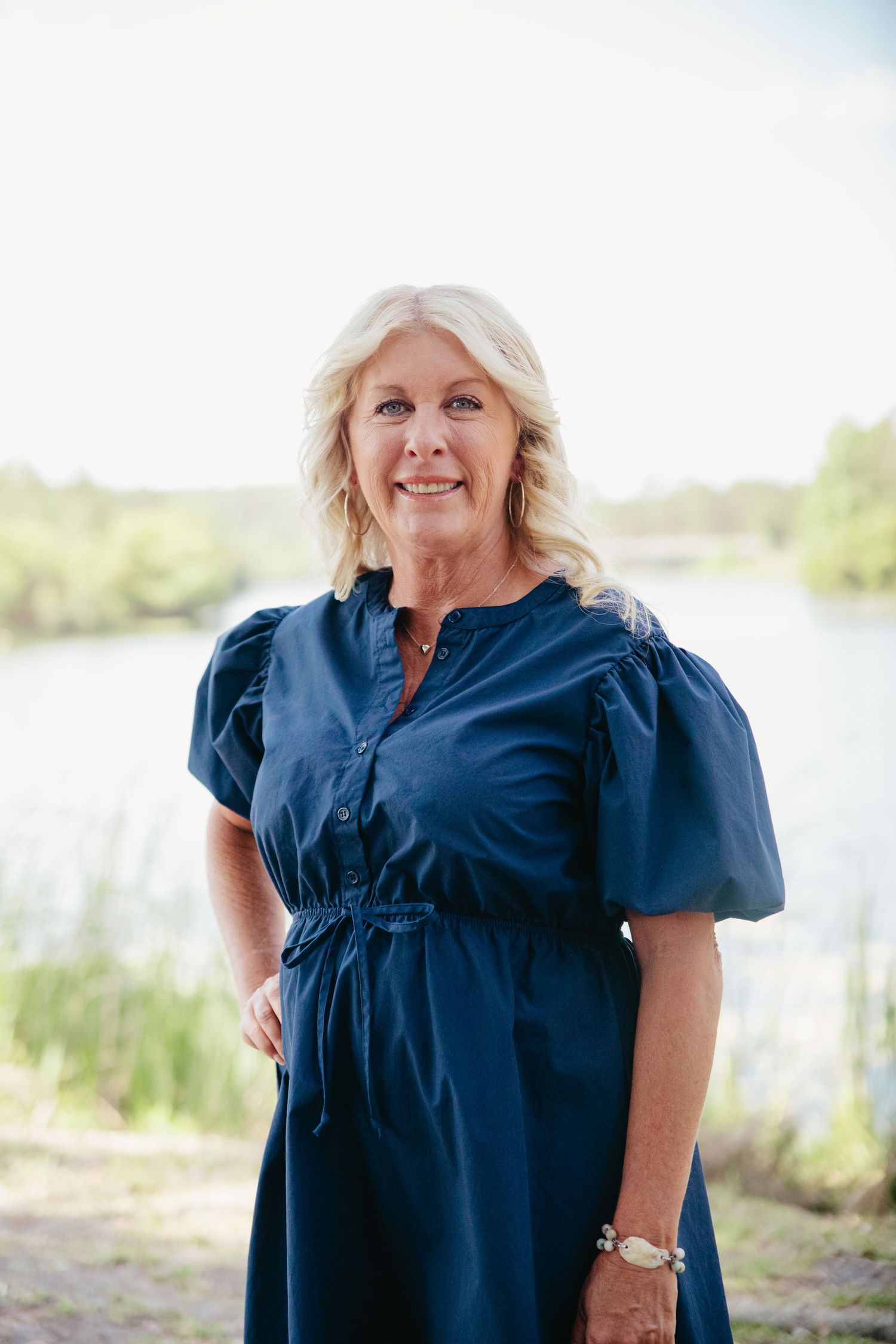 Smiling older blonde woman in navy blue puff-sleeve dress posing outdoors near a peaceful pond.