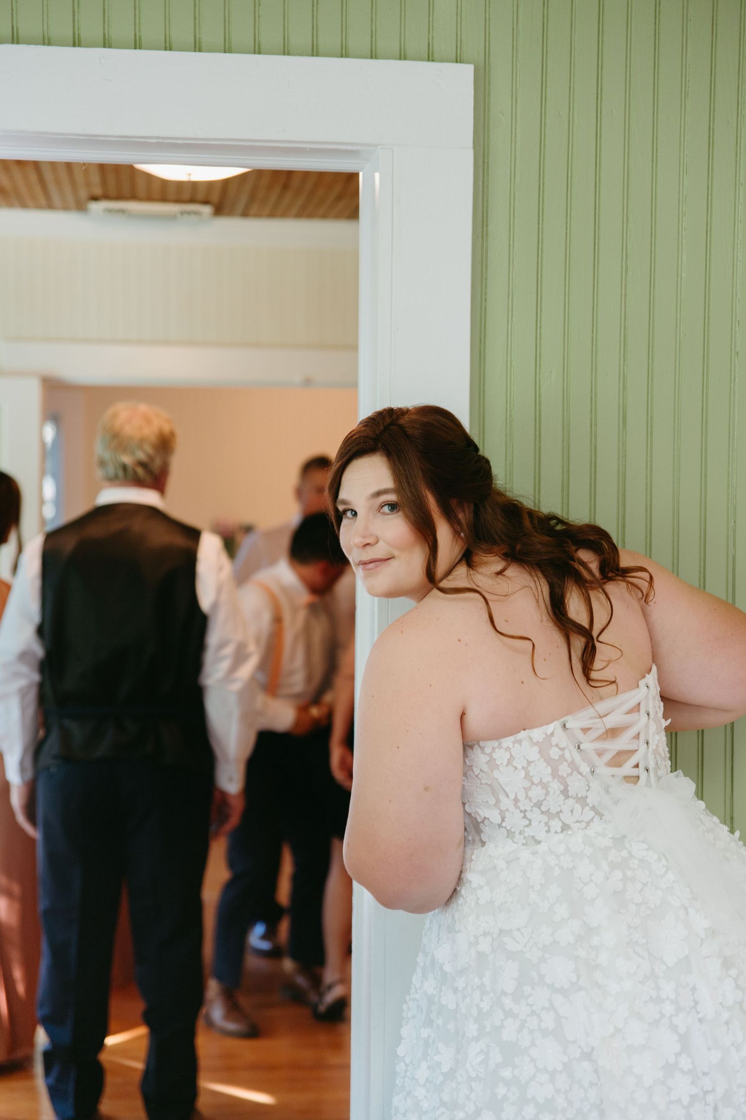Bride in strapless floral wedding dress smiling over her shoulder in a green-walled venue doorway.