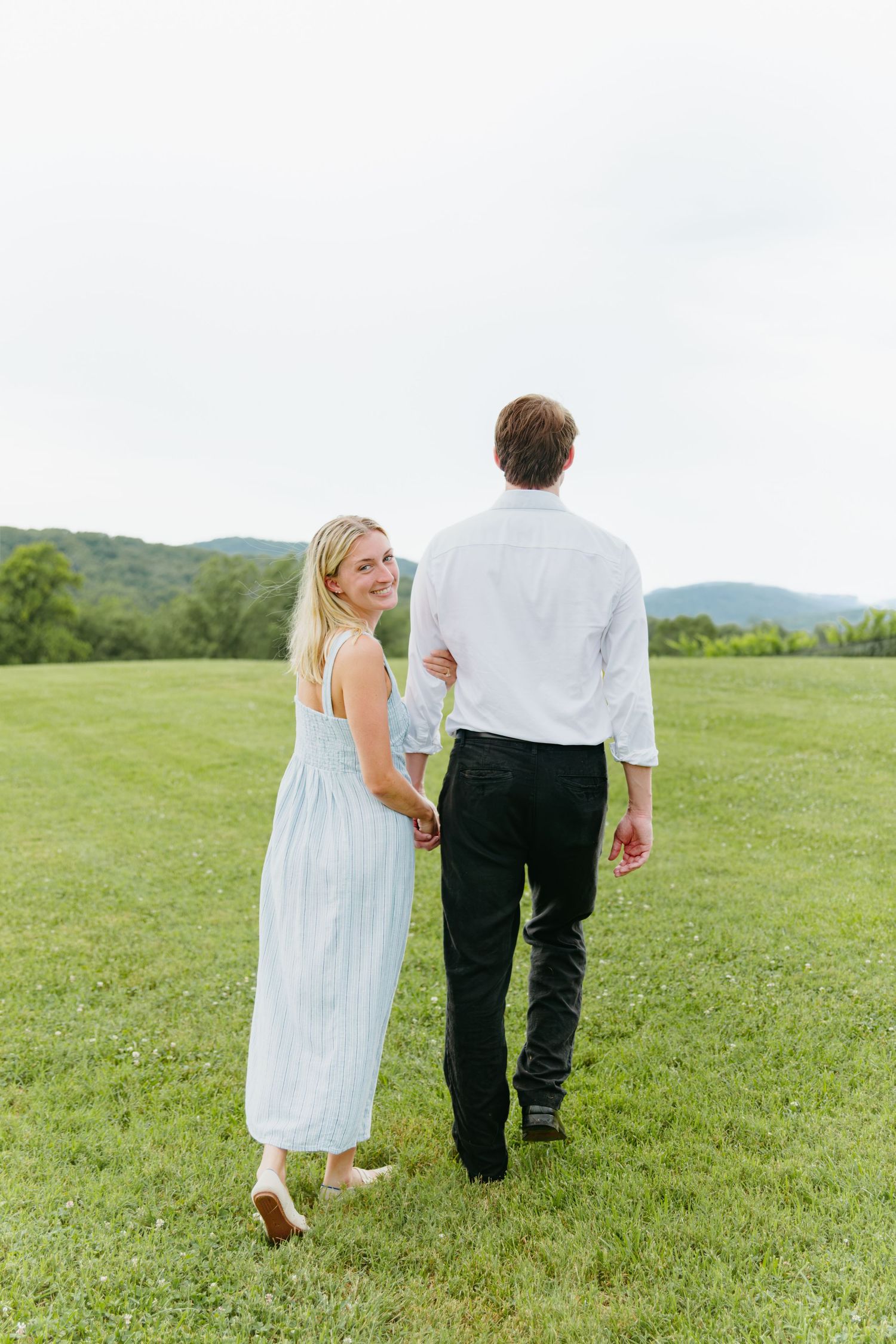 Newly engaged couple walks hand in hand across a green meadow with mountain views, woman smiling back at camera.