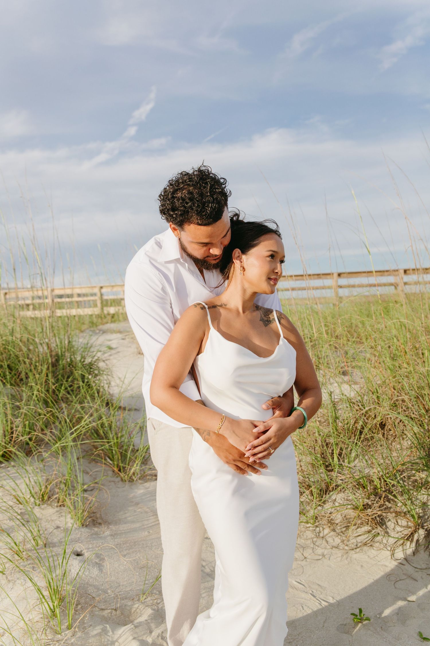 Couple in white embracing on sandy beach dunes with sea oats and blue sky.