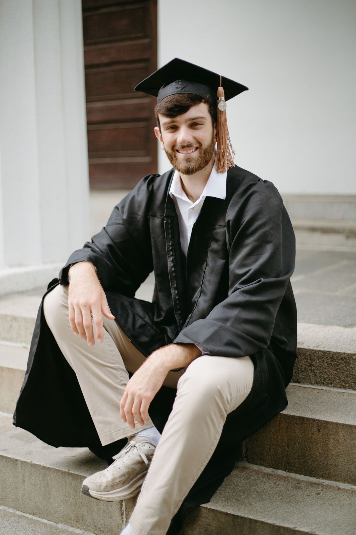 Smiling young man in graduation cap and gown sits on stone steps outside a building.